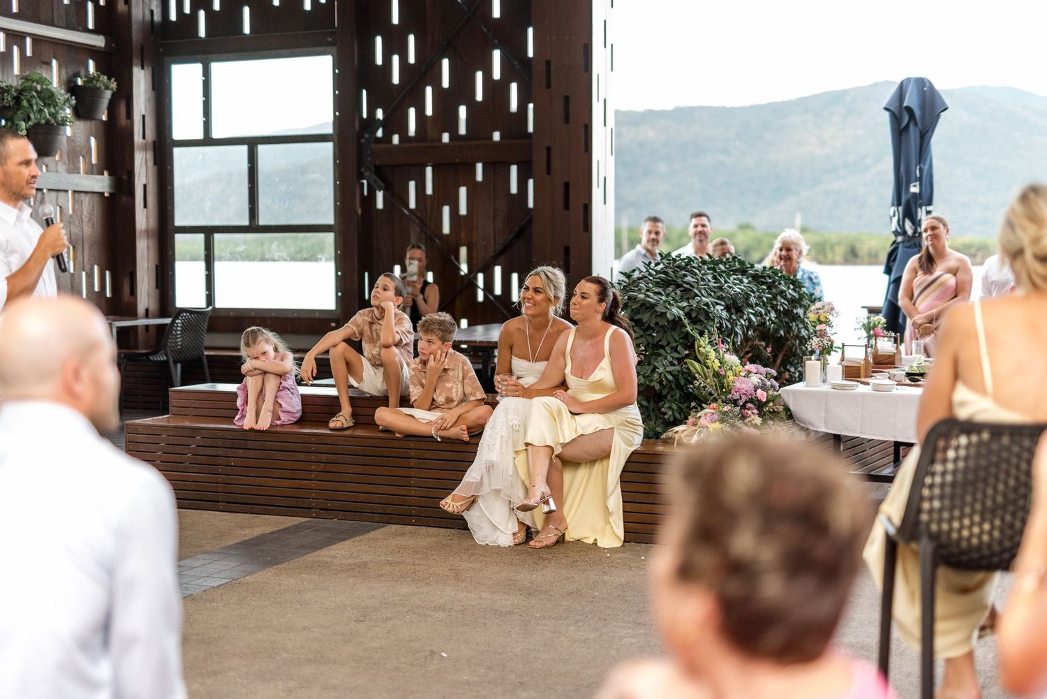 Wedding guests seated on wooden benches at modern venue with mountain views and decorative wall.