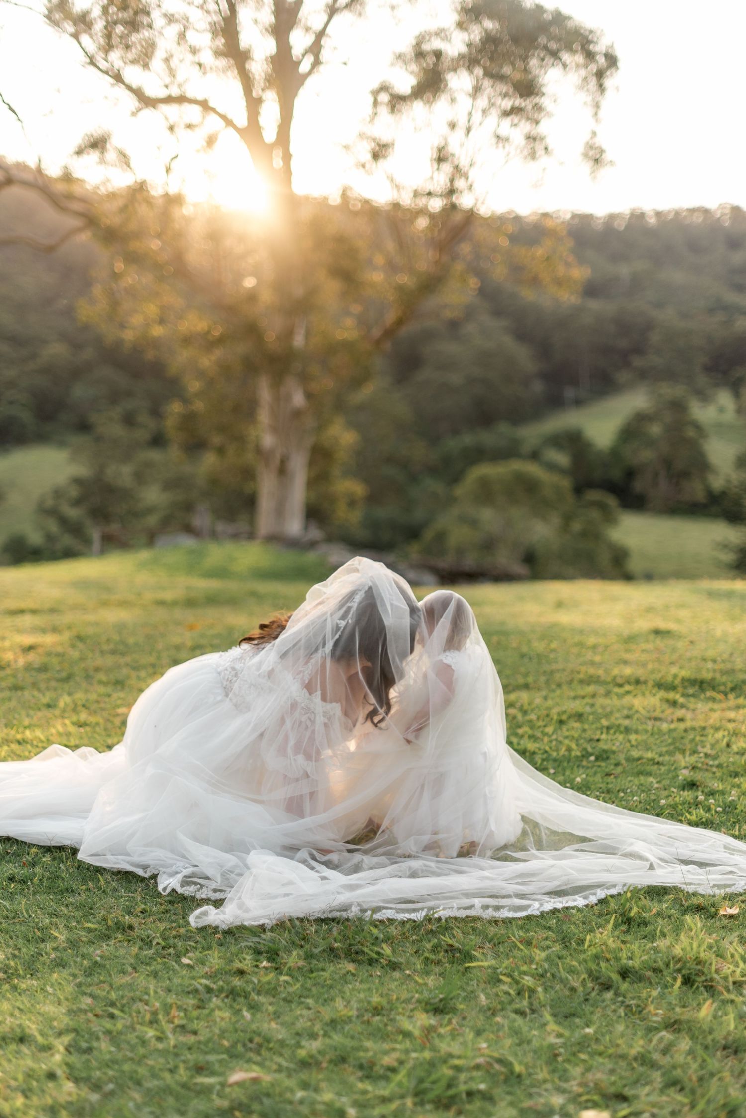 Bride and groom kissing under flowing veil while seated on sunlit green hillside.