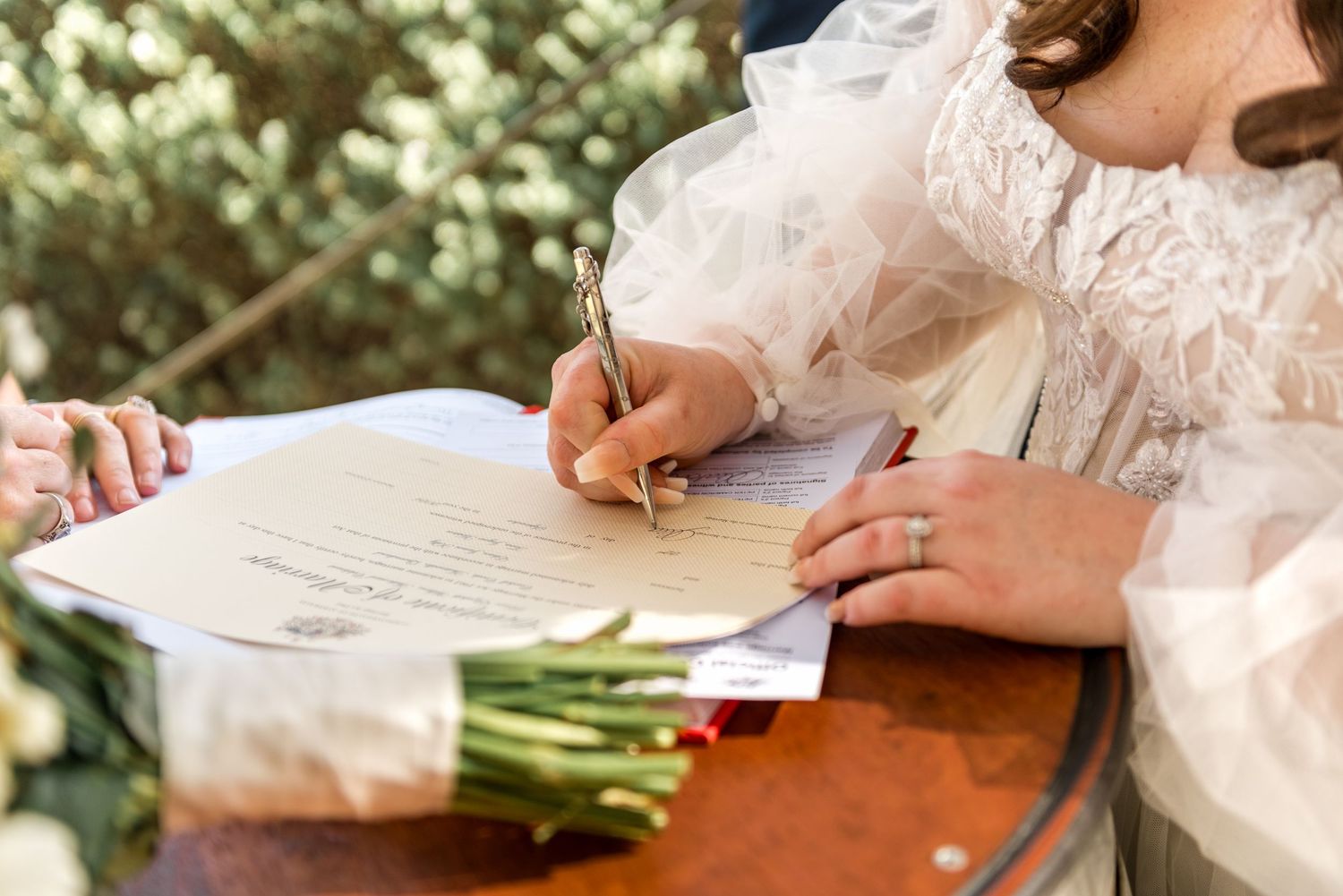 Bride signing wedding certificate with gold pen, wearing elegant white lace gown, with floral bouquet on wooden table.