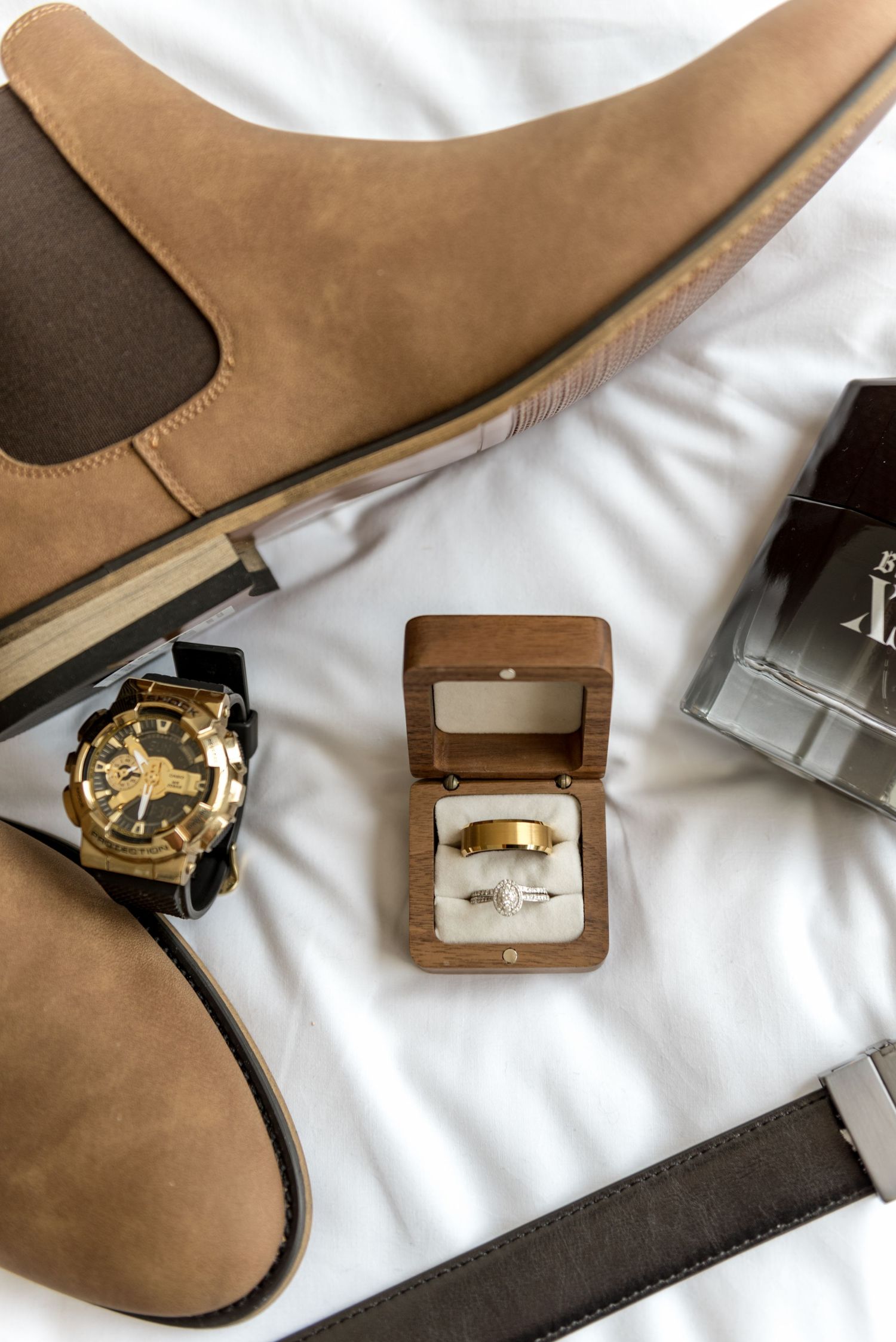 Wedding flat lay with gold rings in wooden box, suede Chelsea boots, gold watch, and cologne.