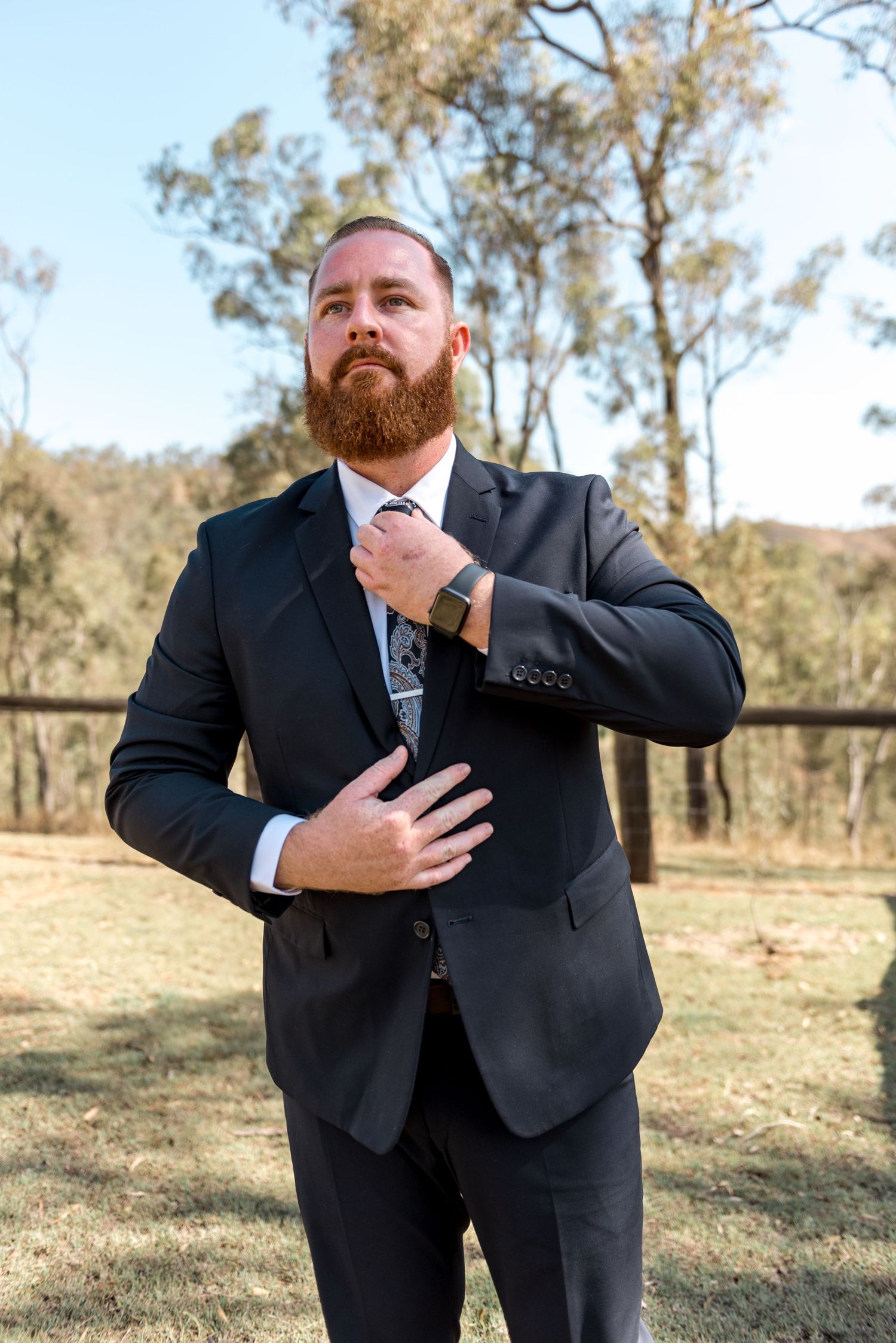 Bearded groom in black suit adjusting tie outdoors at rustic wedding venue with eucalyptus trees.