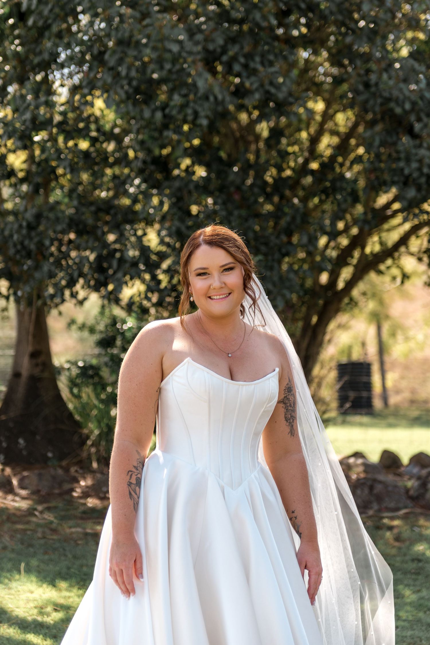 Radiant plus-size bride smiles confidently in strapless white ballgown with veil among green trees.