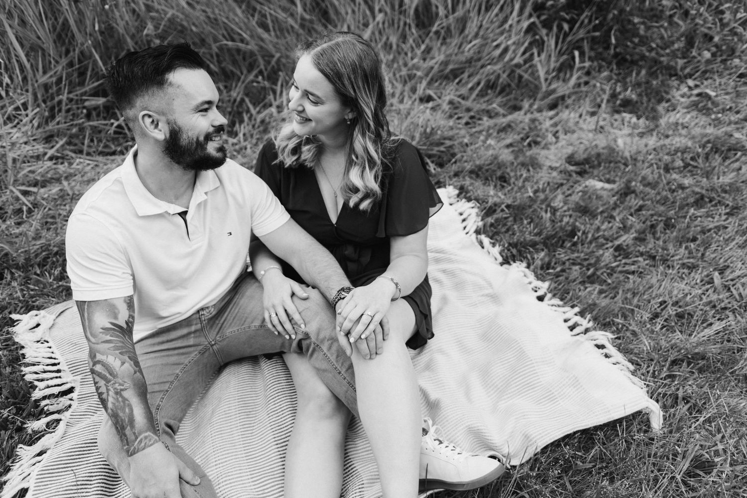 Black and white photo of a couple sitting on a blanket in a field, smiling at each other.