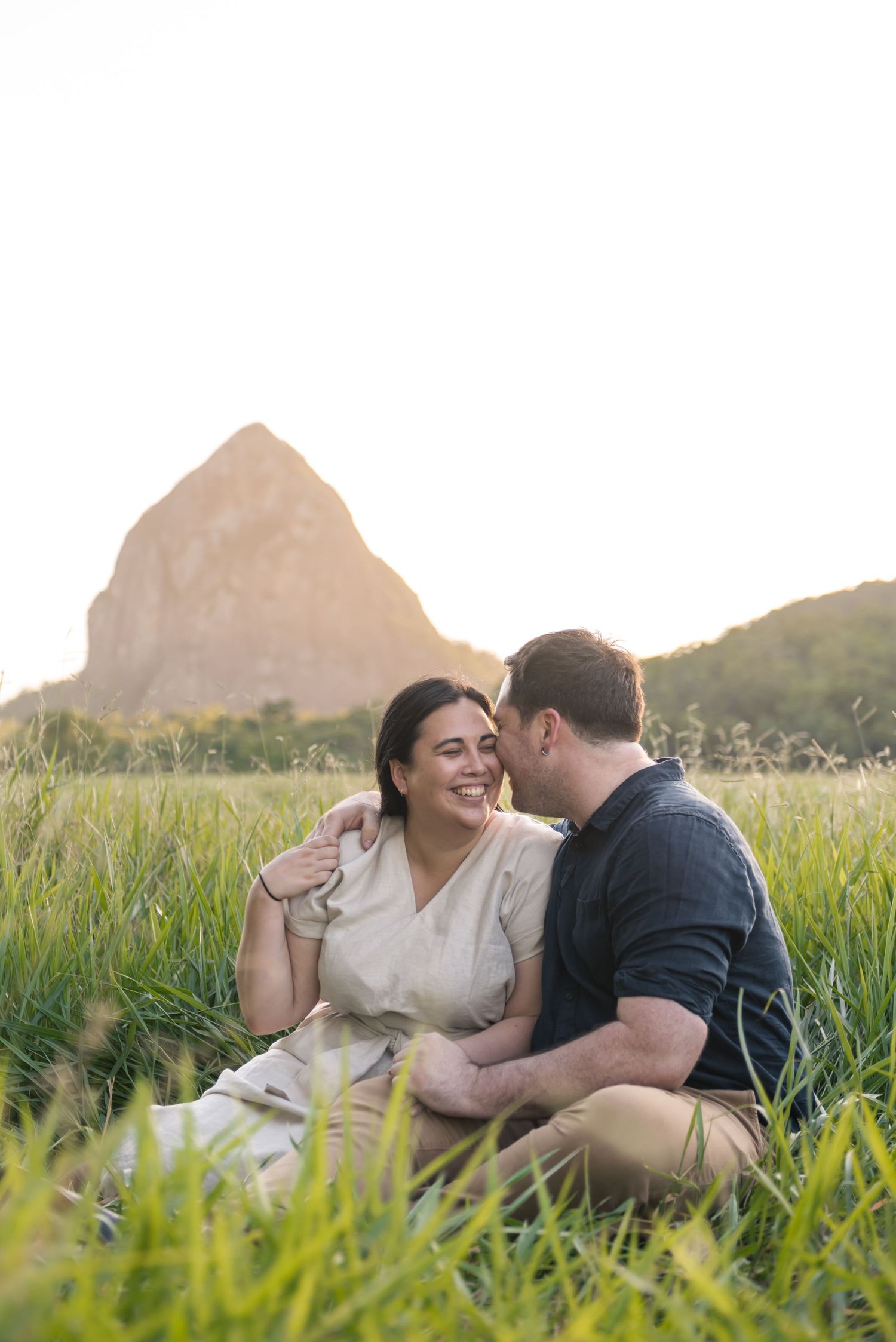 Couple sitting in tall grass laughing together with rocky mountain in background.