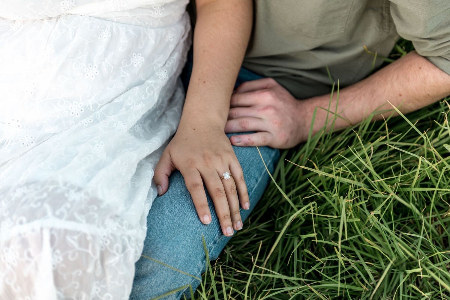 Close-up of couple sitting in green grass, woman wearing engagement ring in white lace dress and mans hand on knee.