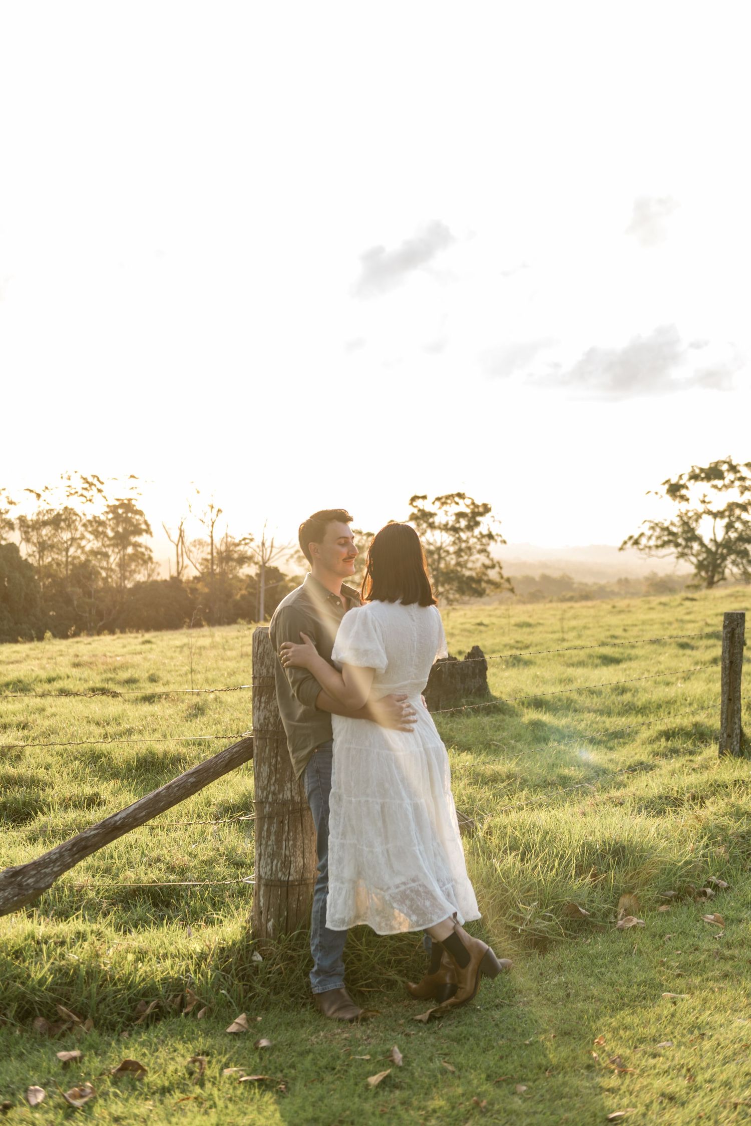 Romantic couple sharing a tender moment at sunset beside a wooden fence in a meadow.
