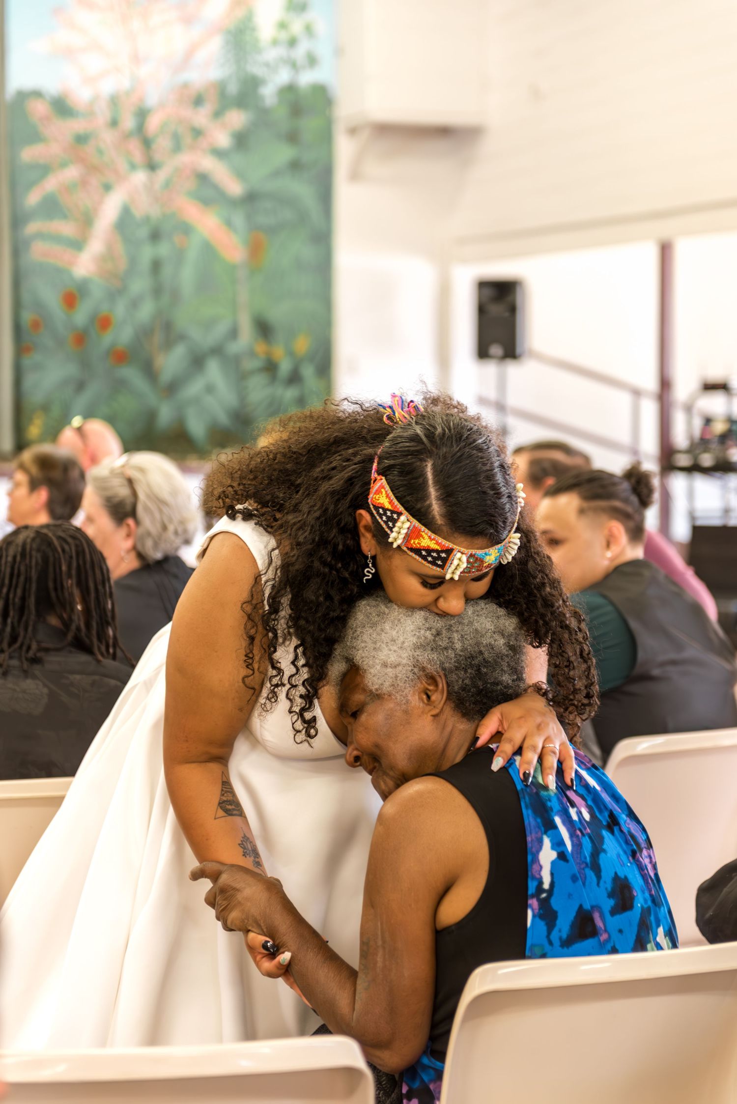 A bride in a white dress embraces an elderly woman at a wedding ceremony indoors.