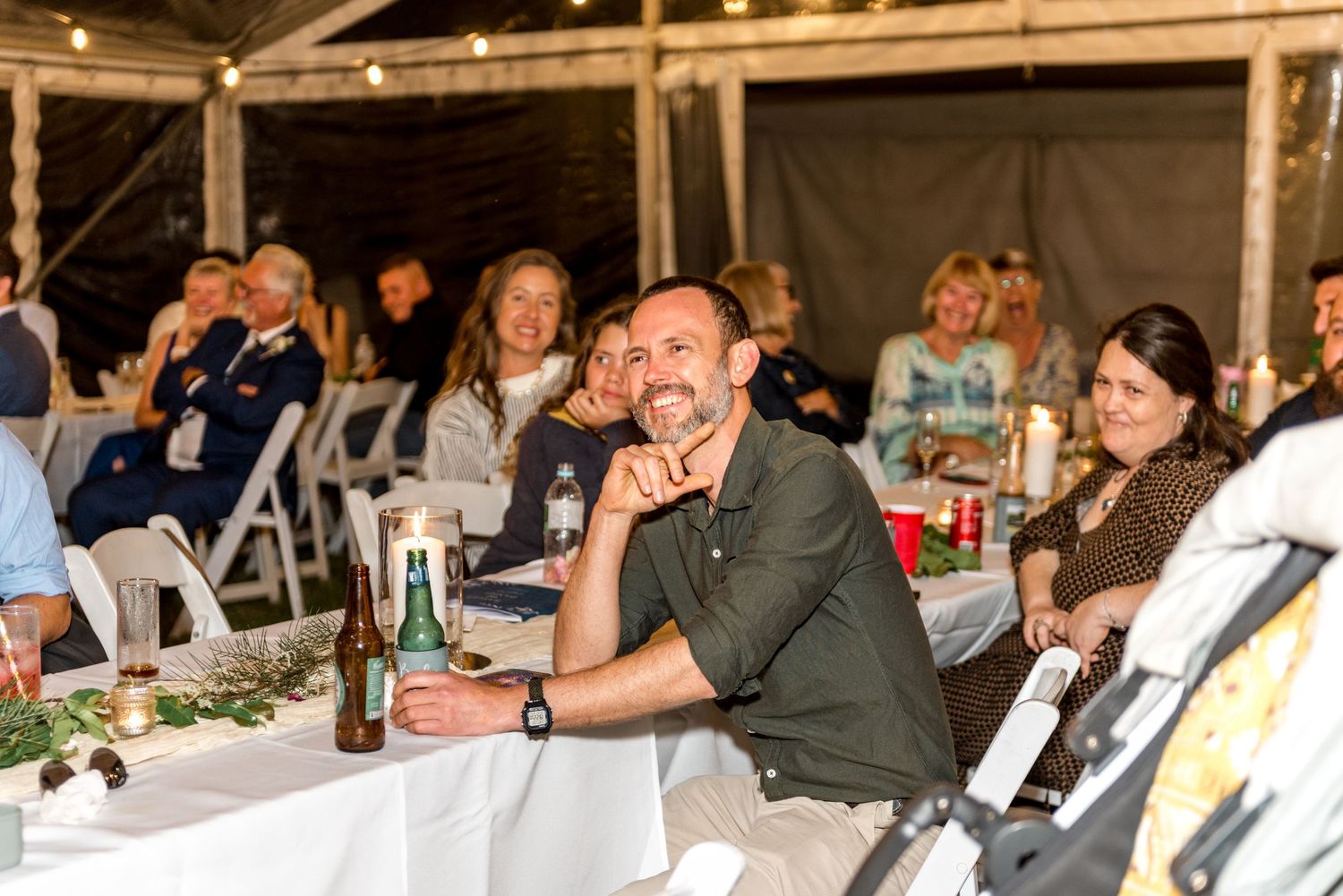 Smiling bearded man in green shirt enjoys drinks with guests at a festive outdoor wedding reception under a tent.