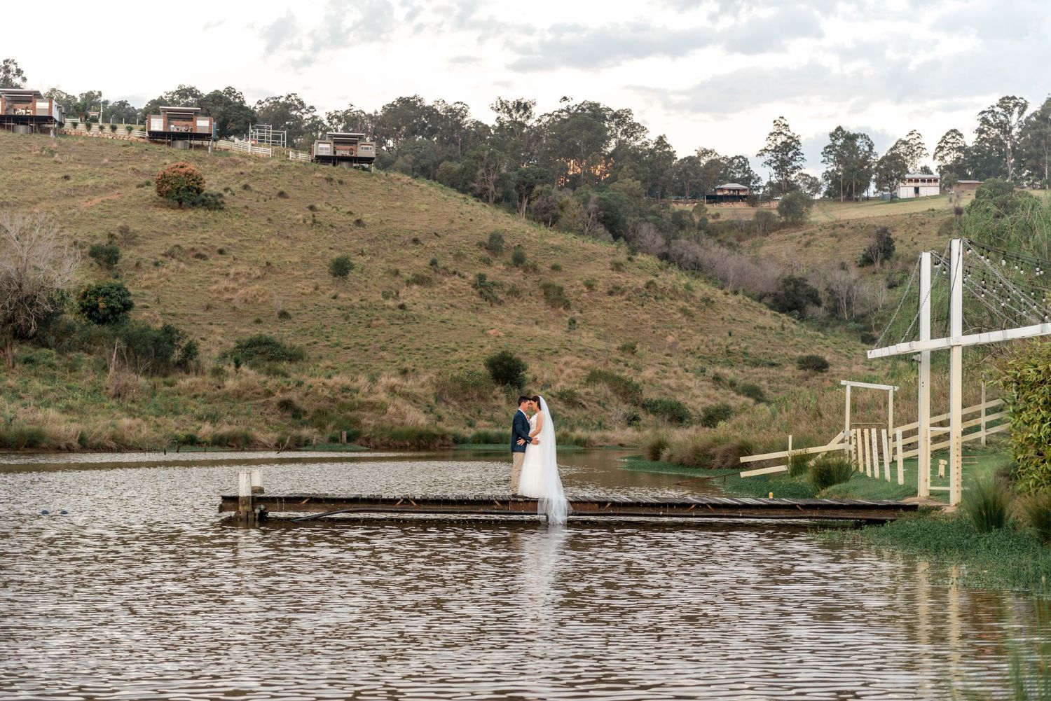 Bride and groom share a romantic embrace on a wooden dock surrounded by a serene lake and rolling hills.