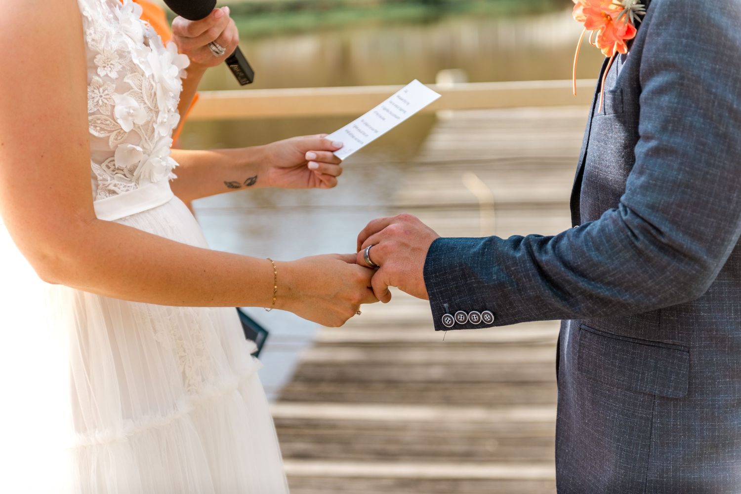 Bride and groom hold hands during vow exchange at outdoor wedding ceremony with floral decorations nearby.