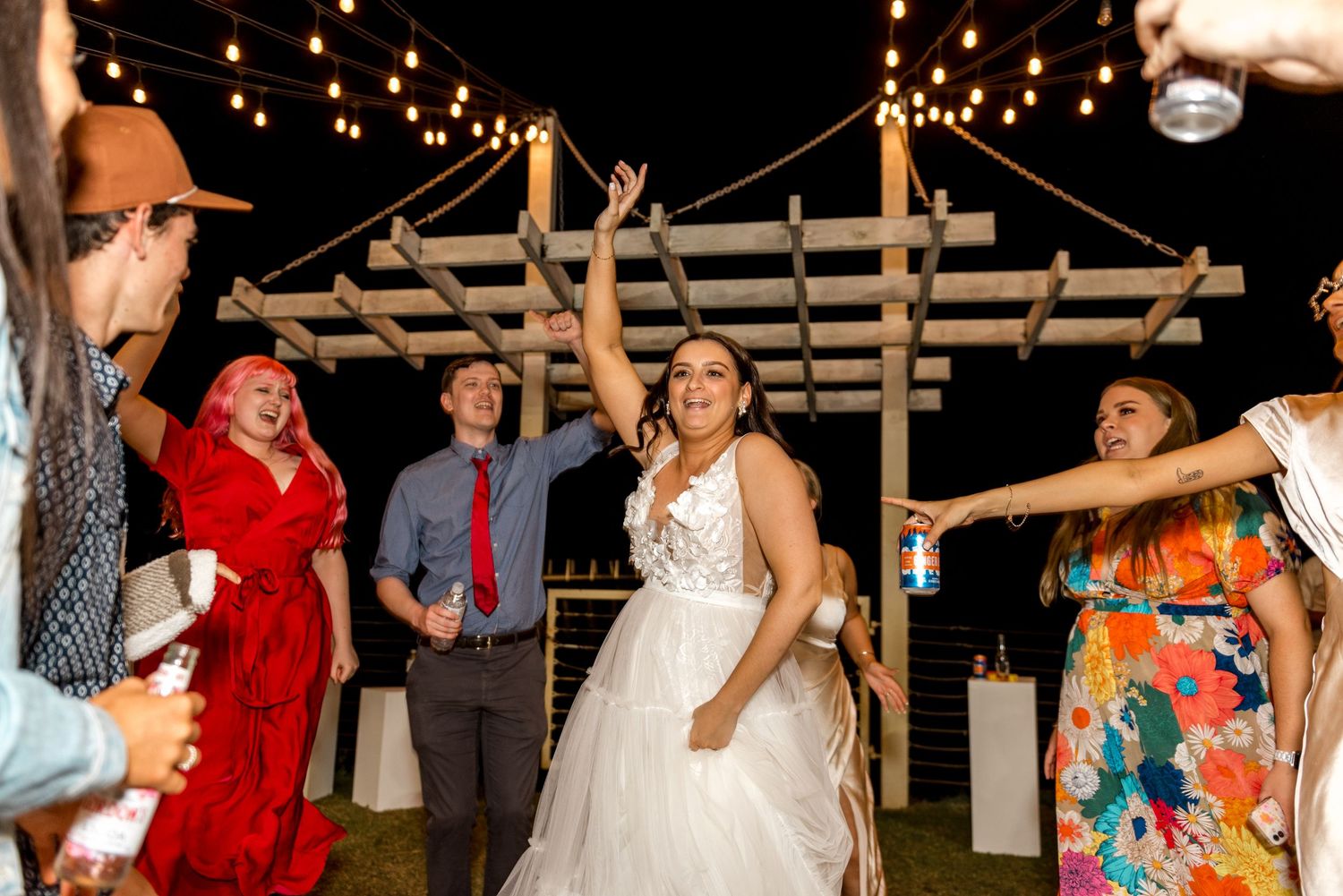 Bride in white ball gown dancing joyfully with guests at outdoor wedding reception under string lights.