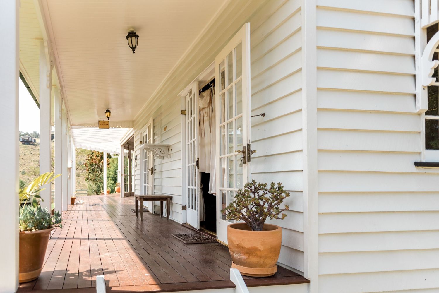 Elegant white weatherboard farmhouse verandah with potted plants and timber decking.