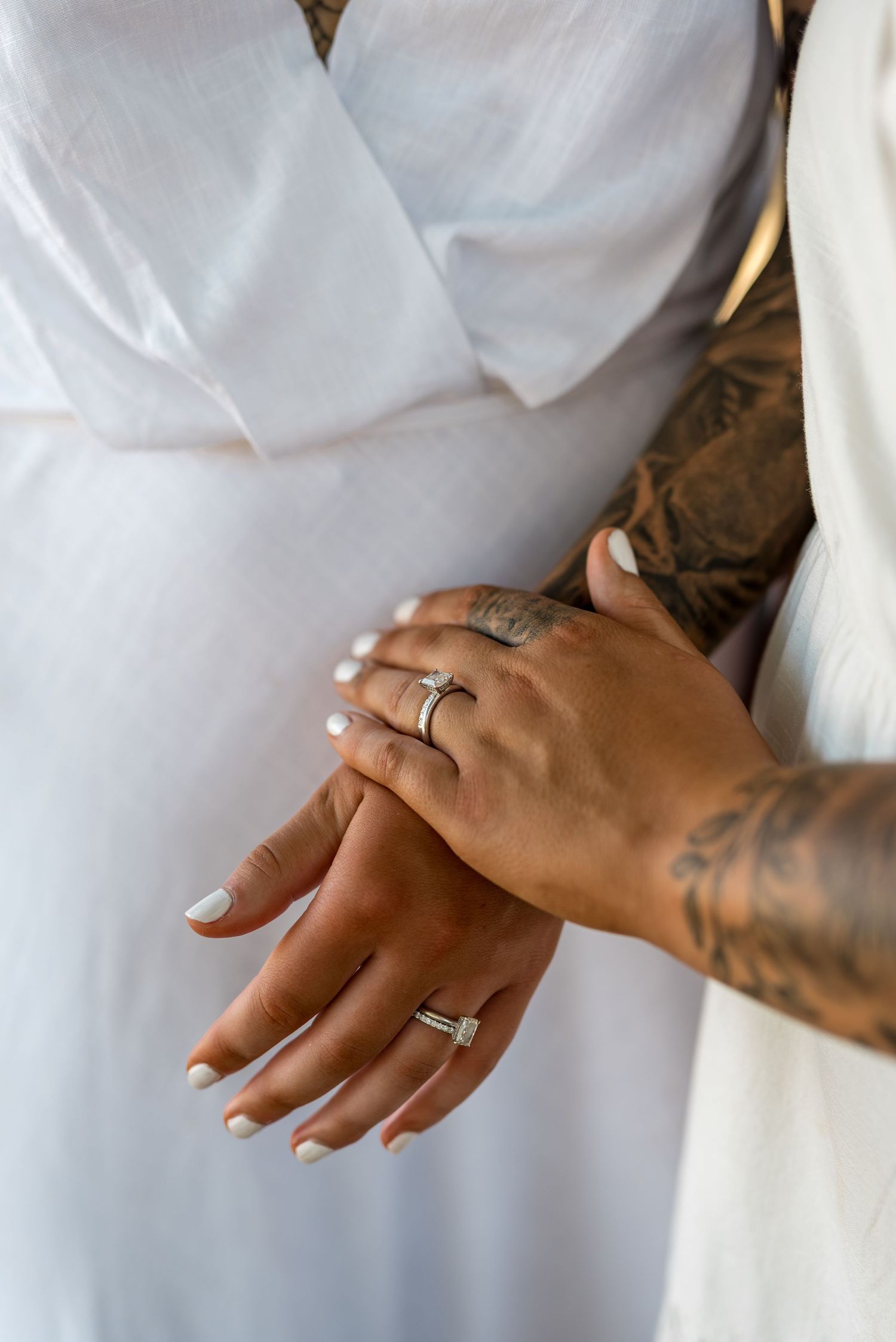 Close-up of two hands with engagement rings and tattoos clasped together against a white dress fabric.