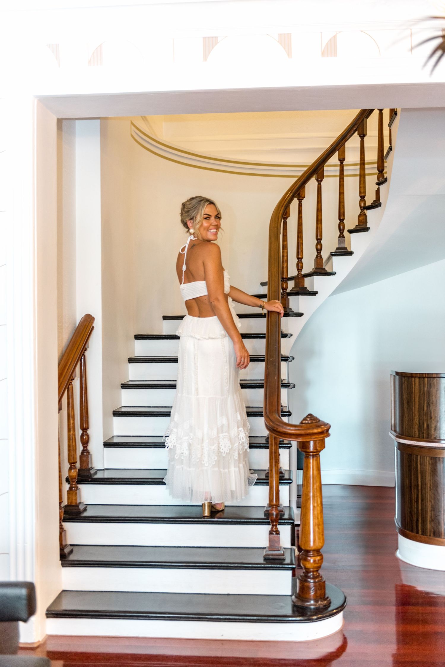 Bride in white two-piece wedding dress poses on elegant curved staircase with dark wood railings.