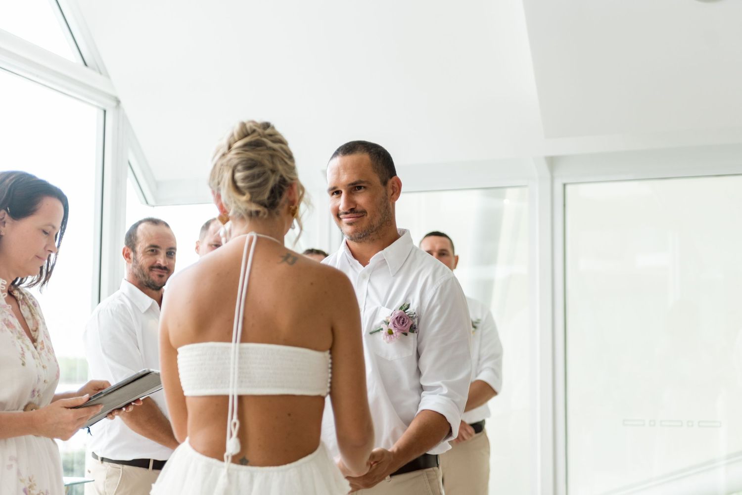 Groom smiling at bride during intimate wedding ceremony while officiant reads vows beside them.