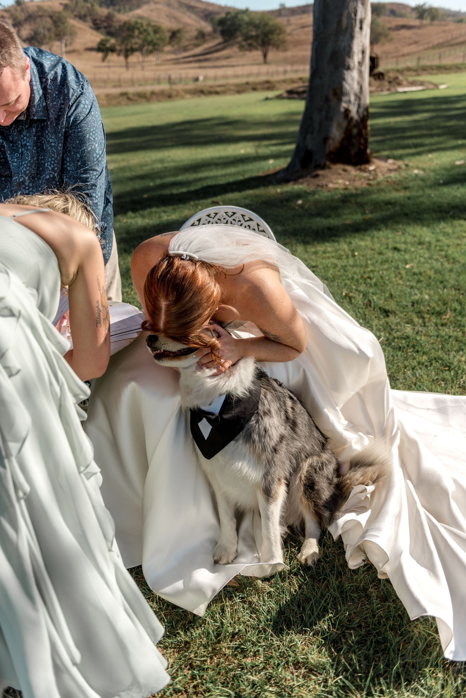 Bride in white gown leans down to nuzzle her dog wearing a tuxedo outfit during outdoor wedding ceremony.