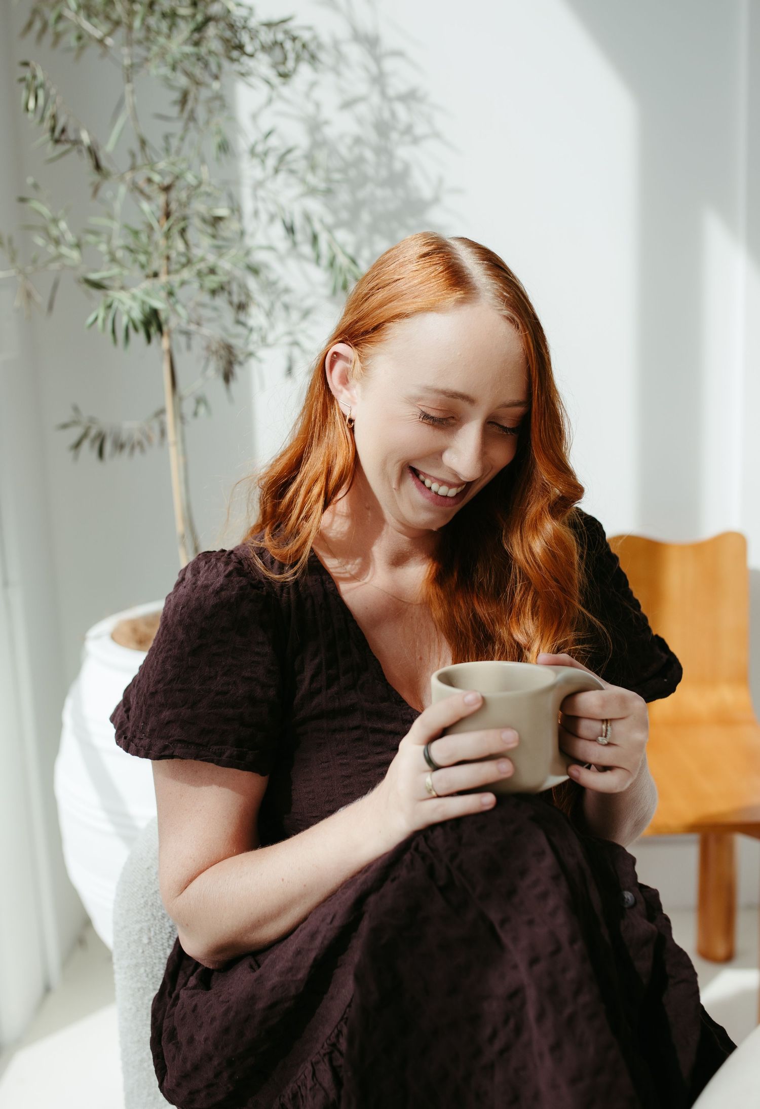Smiling redhead woman in dark dress holding ceramic mug, seated in bright room.