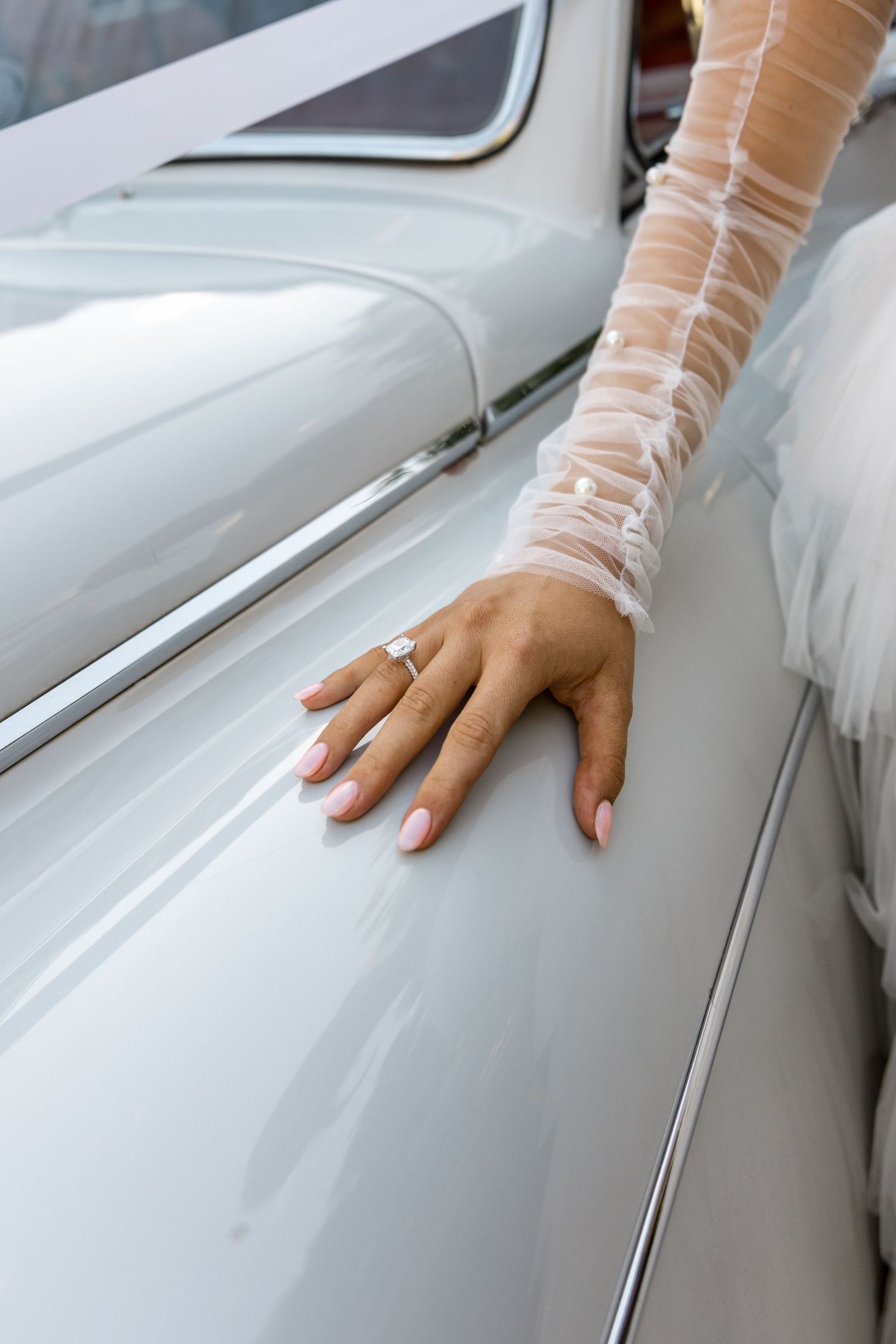 Bride's hand with diamond engagement ring resting on white vintage car, wearing sheer lace wedding glove.
