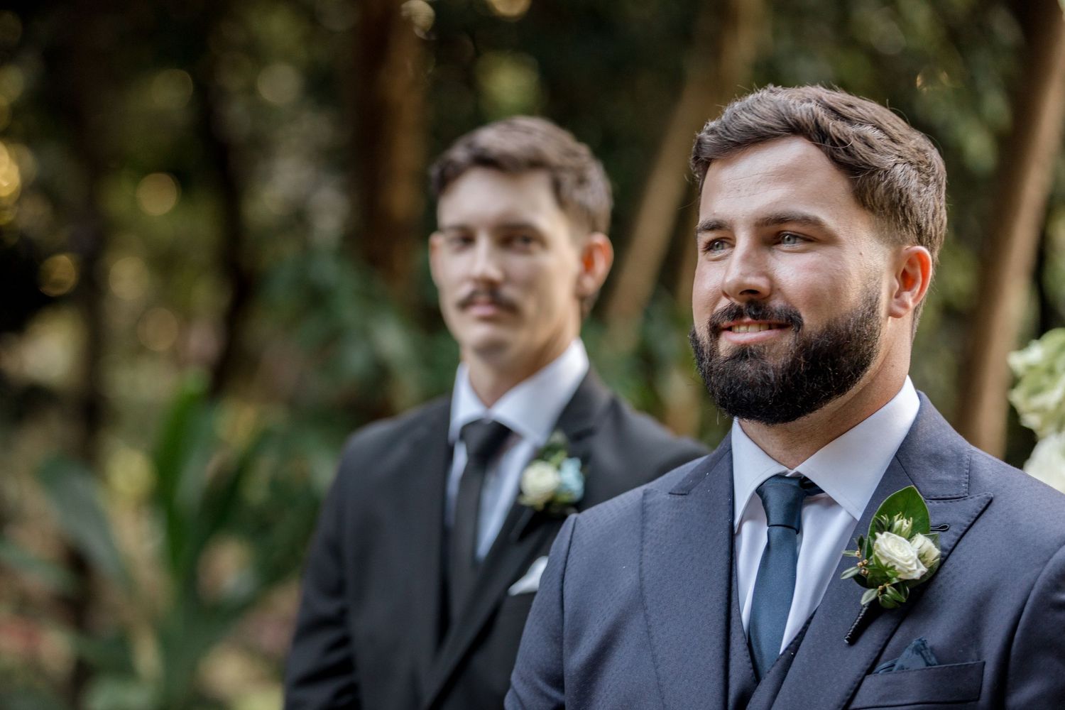 Bearded groom in navy suit watches bride approach at outdoor garden wedding ceremony.