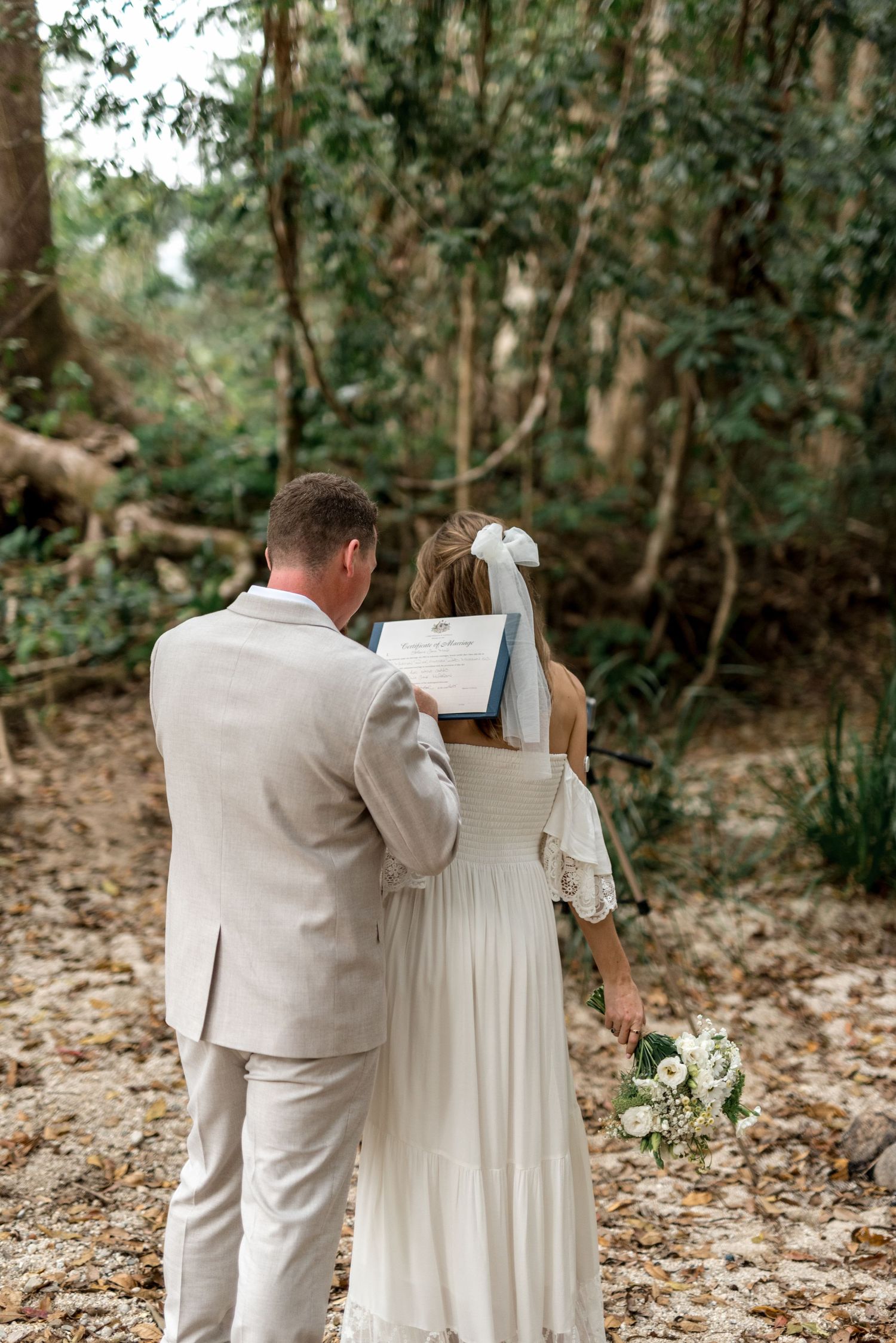 Newlyweds view their marriage certificate together from behind in a lush forest setting.