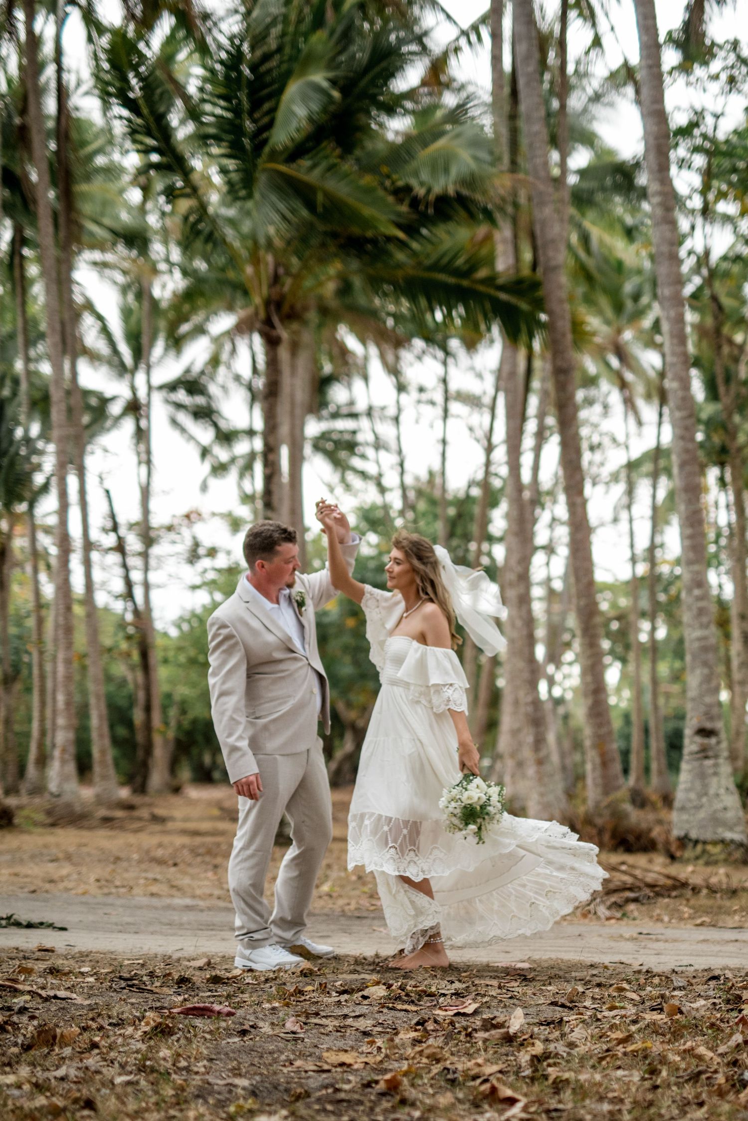 Groom twirls bride in white lace dress among tall palm trees during outdoor tropical wedding shoot.