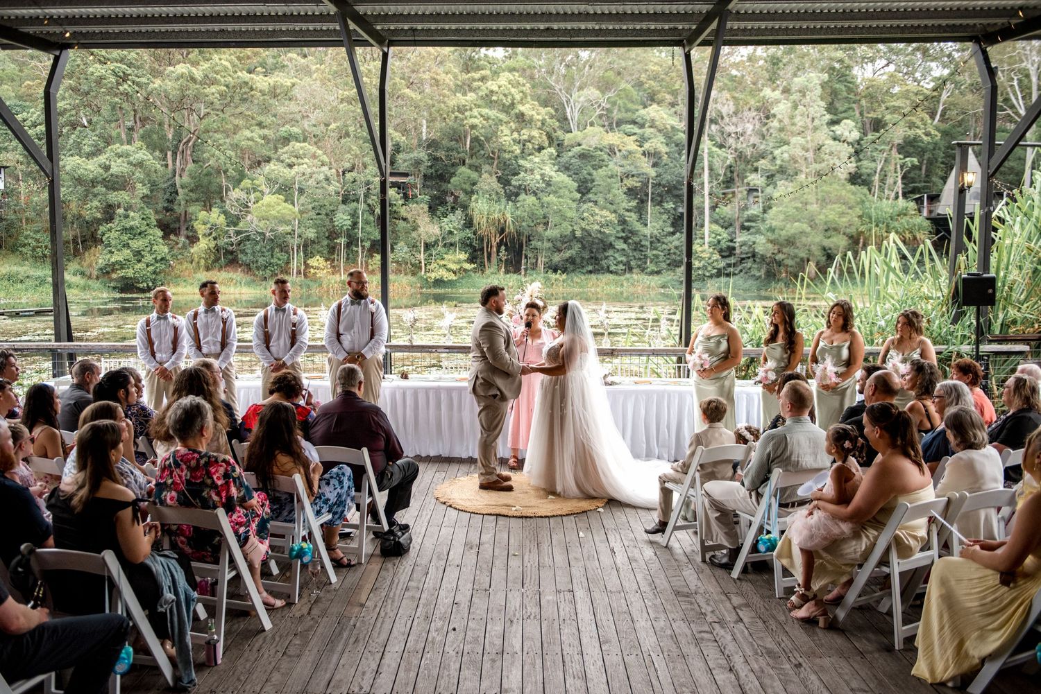 Bride and groom exchange vows at outdoor pavilion wedding with bridal party and guests.