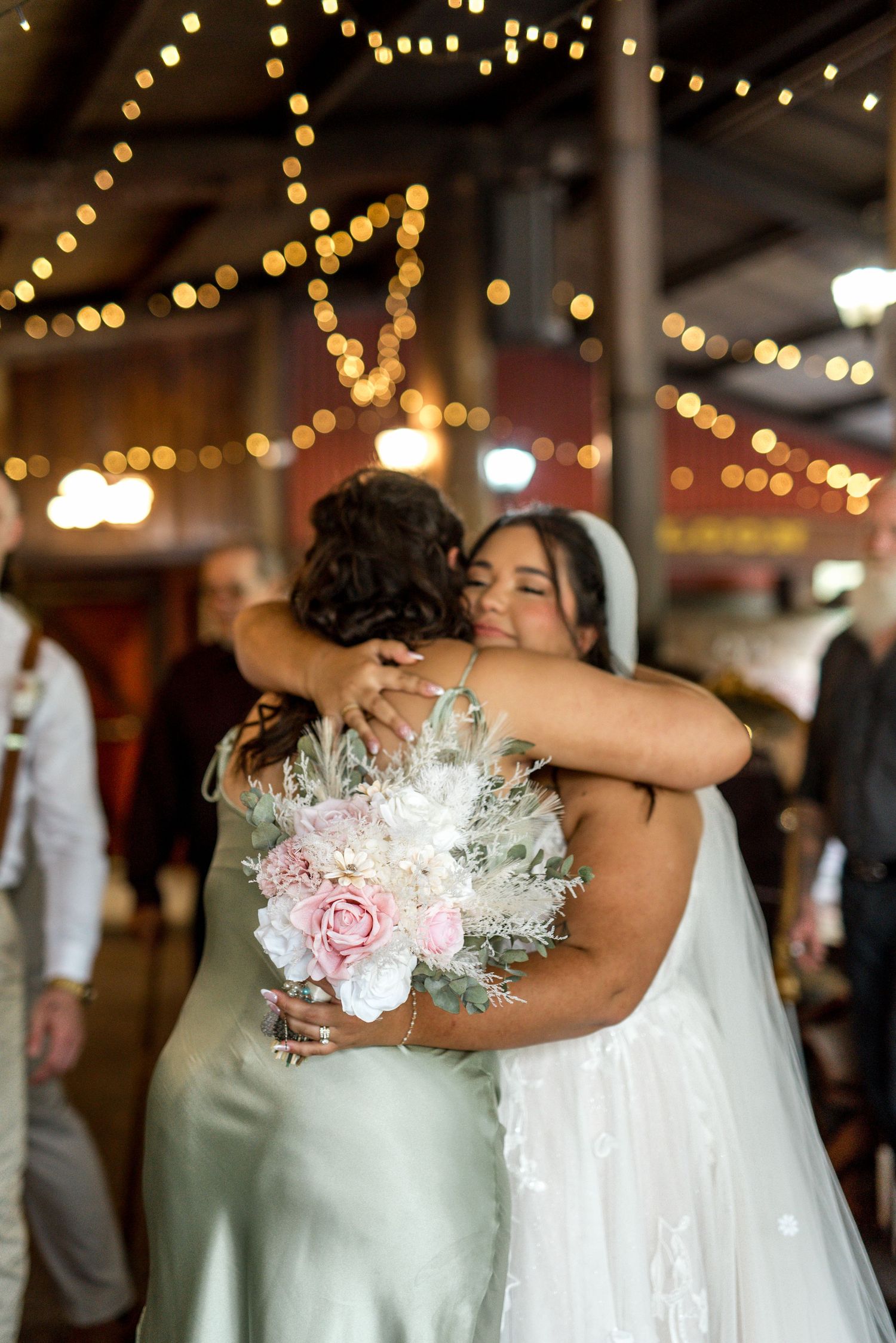 Bride in white gown and veil shares a warm hug with bridesmaid under string lights at reception.