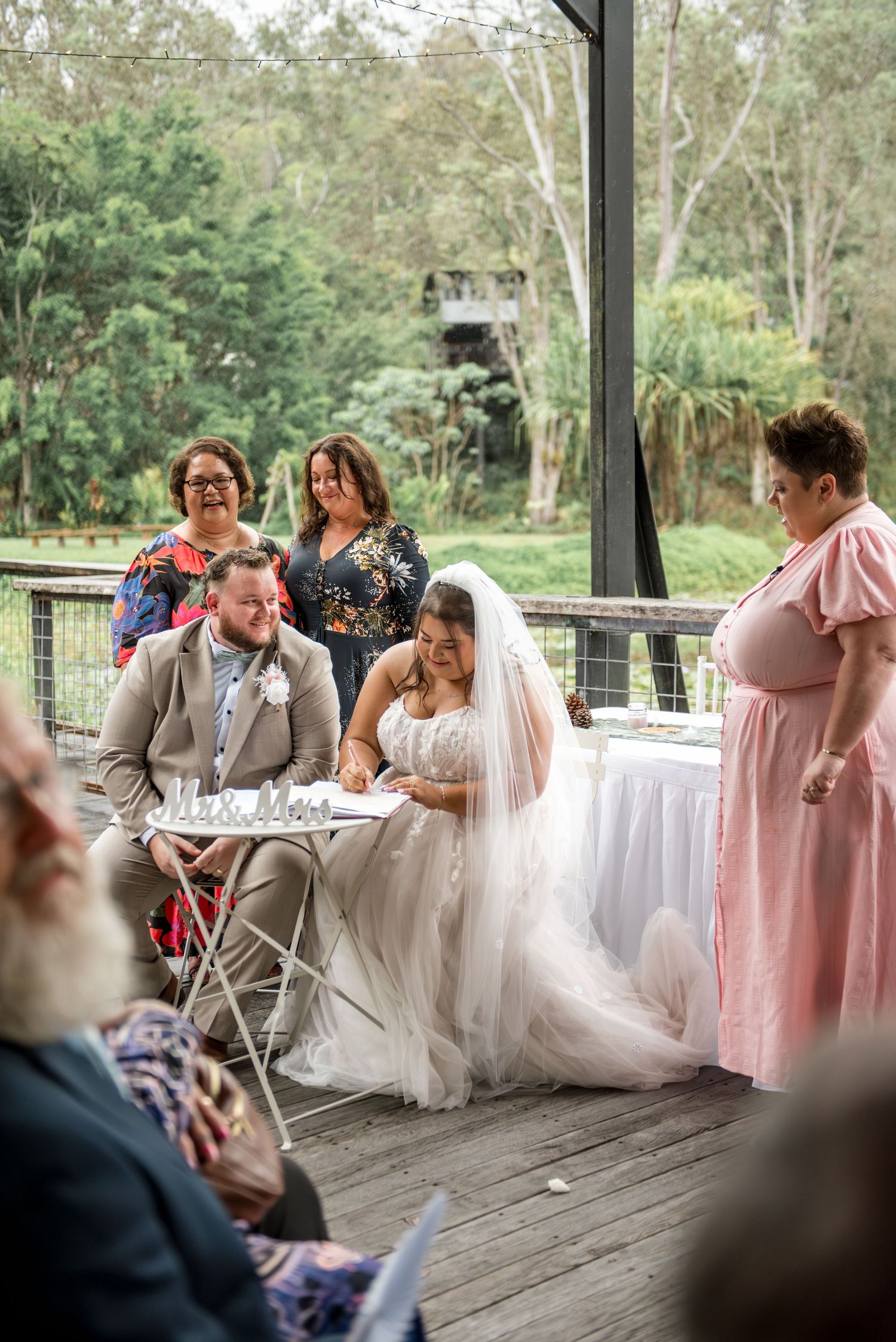 Bride and groom sign marriage certificate at wedding ceremony table with witnesses and guests looking on.