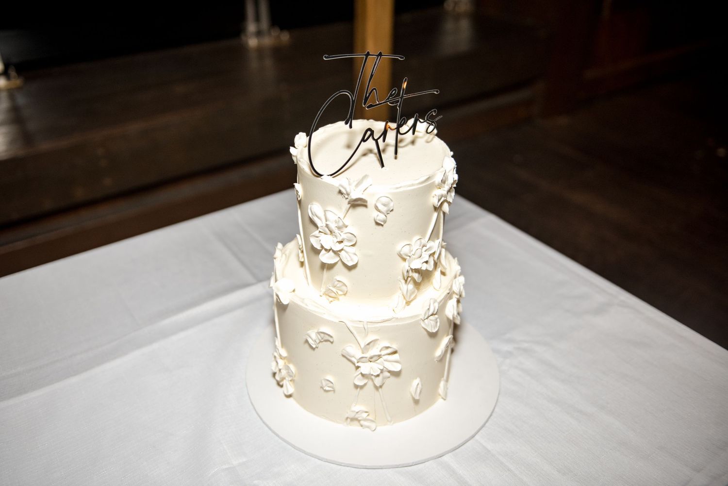 Elegant two-tier white wedding cake decorated with textured sugar flowers and a personalized acrylic cake topper.