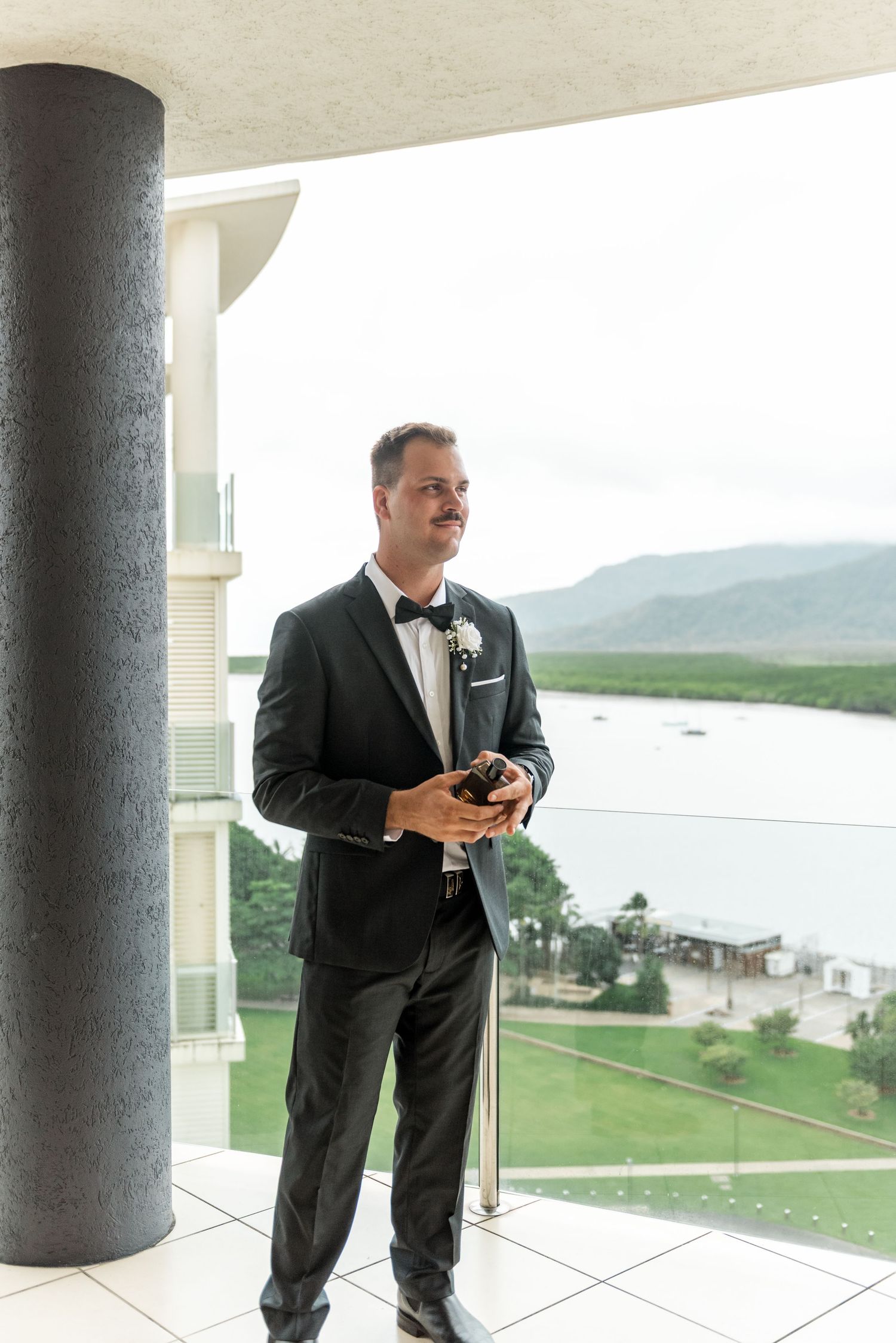 Dapper groom in black tuxedo with boutonniere standing on balcony with scenic water views.