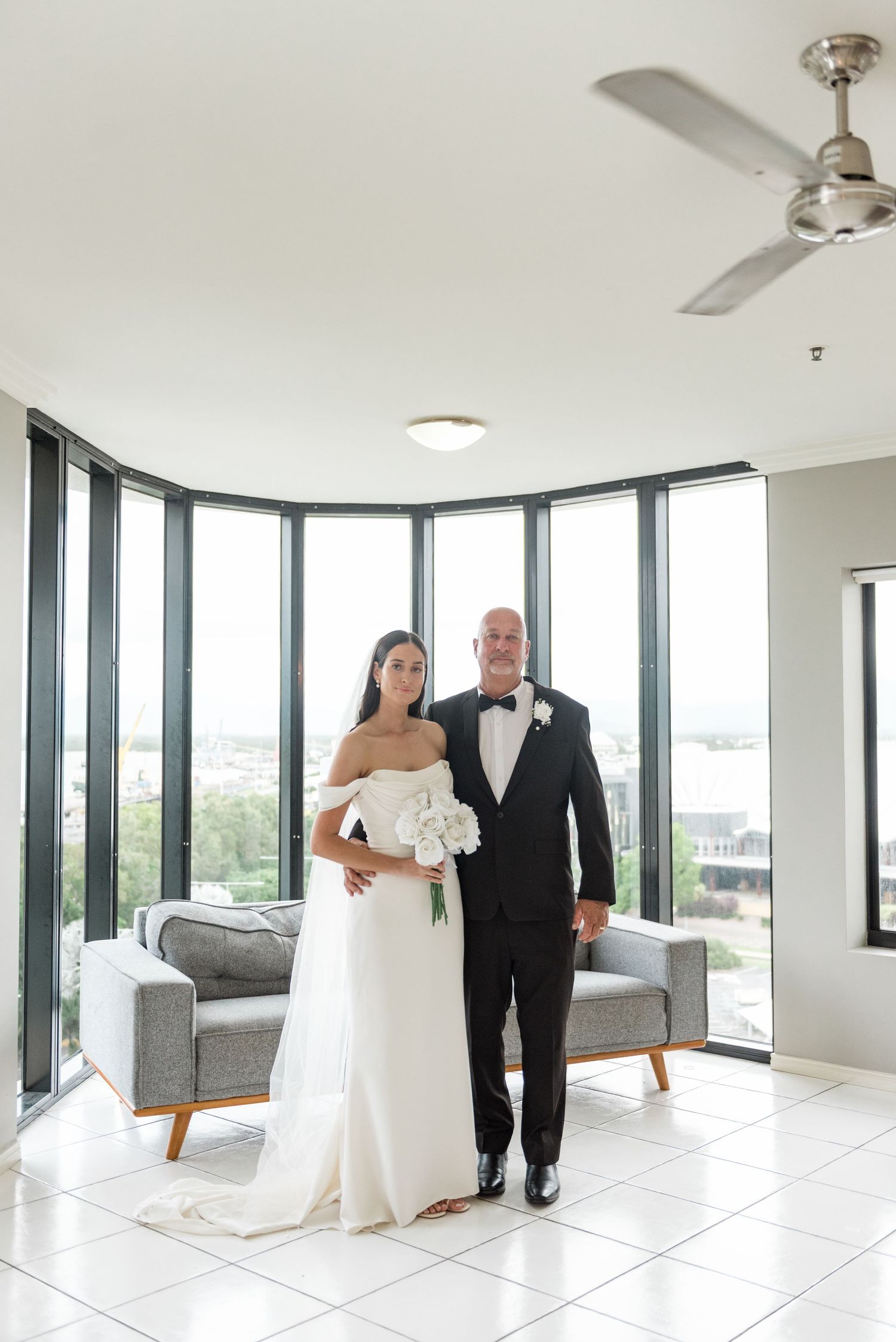 Bride in off-shoulder white gown and groom in black tuxedo pose together in a bright modern room with panoramic windows.