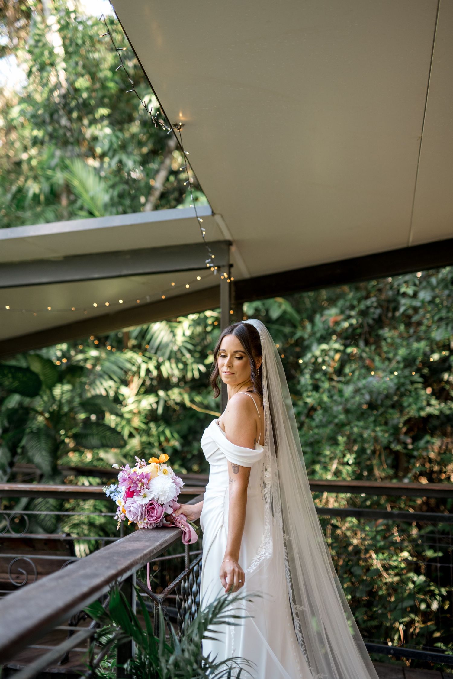 Elegant bride in off-shoulder gown and long veil holds colorful bouquet on a balcony amid lush forest.
