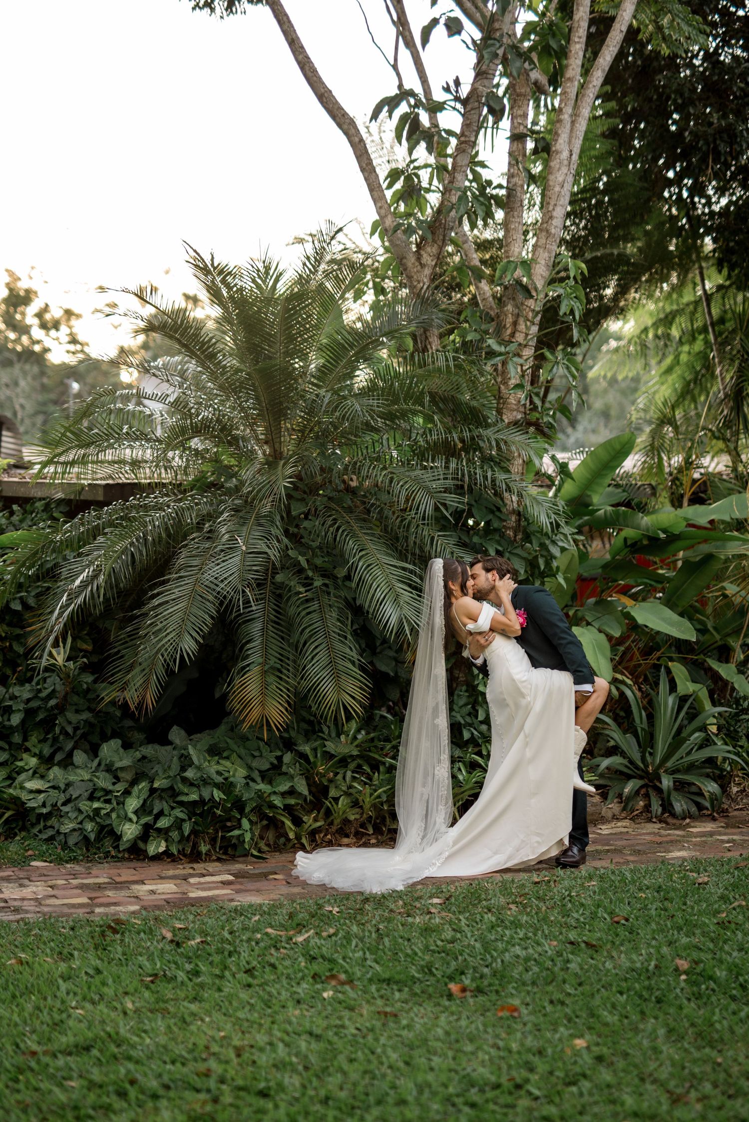 Groom dips bride for a kiss in a lush green garden with palm trees.
