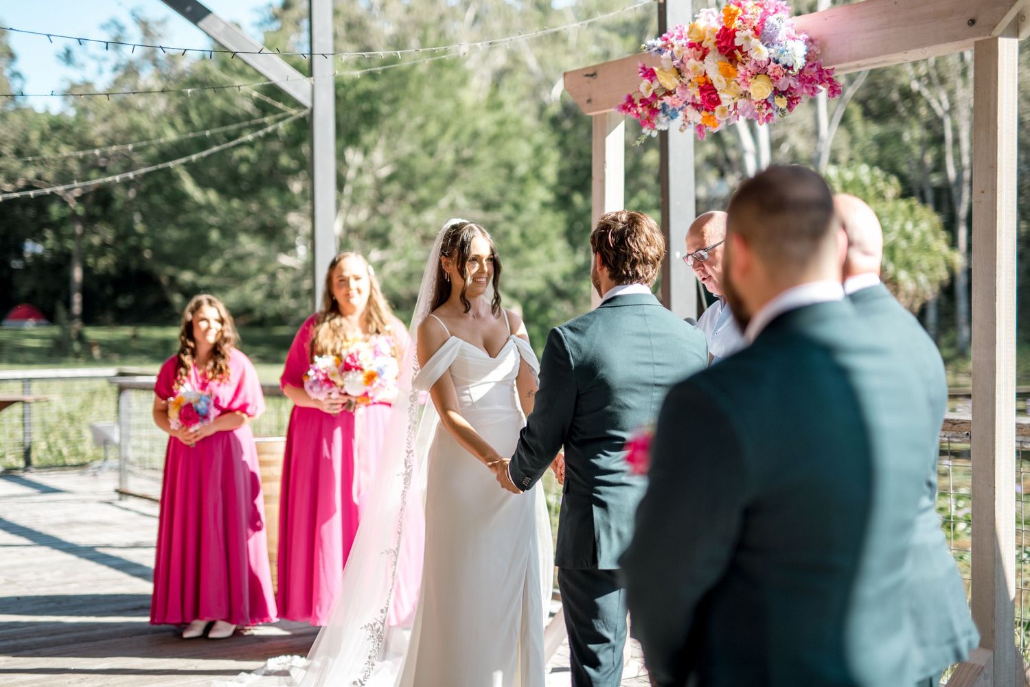 Bride and groom holding hands at outdoor wedding ceremony with bridesmaids in pink dresses and colorful floral arch overhead.