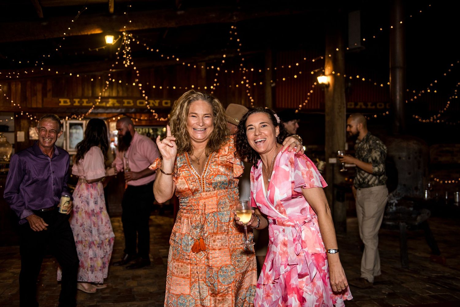Two smiling women in floral dresses celebrating at a lively indoor wedding reception.