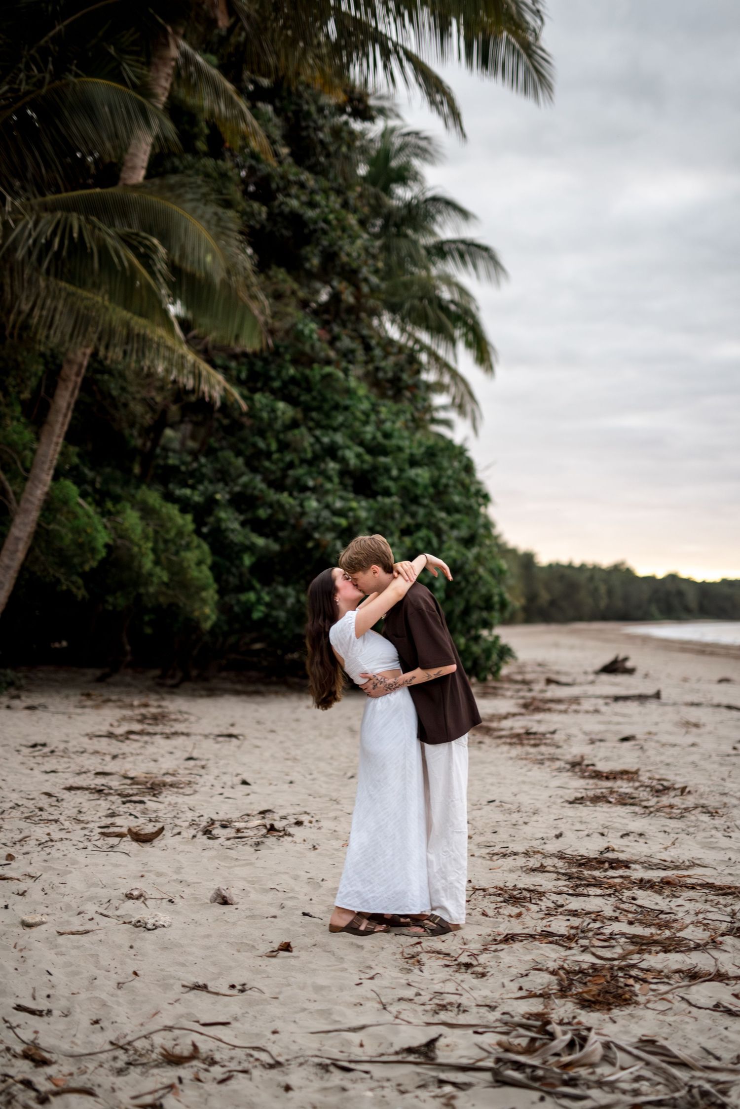Couple sharing a romantic kiss on a tropical beach with tall palm trees and cloudy sky.