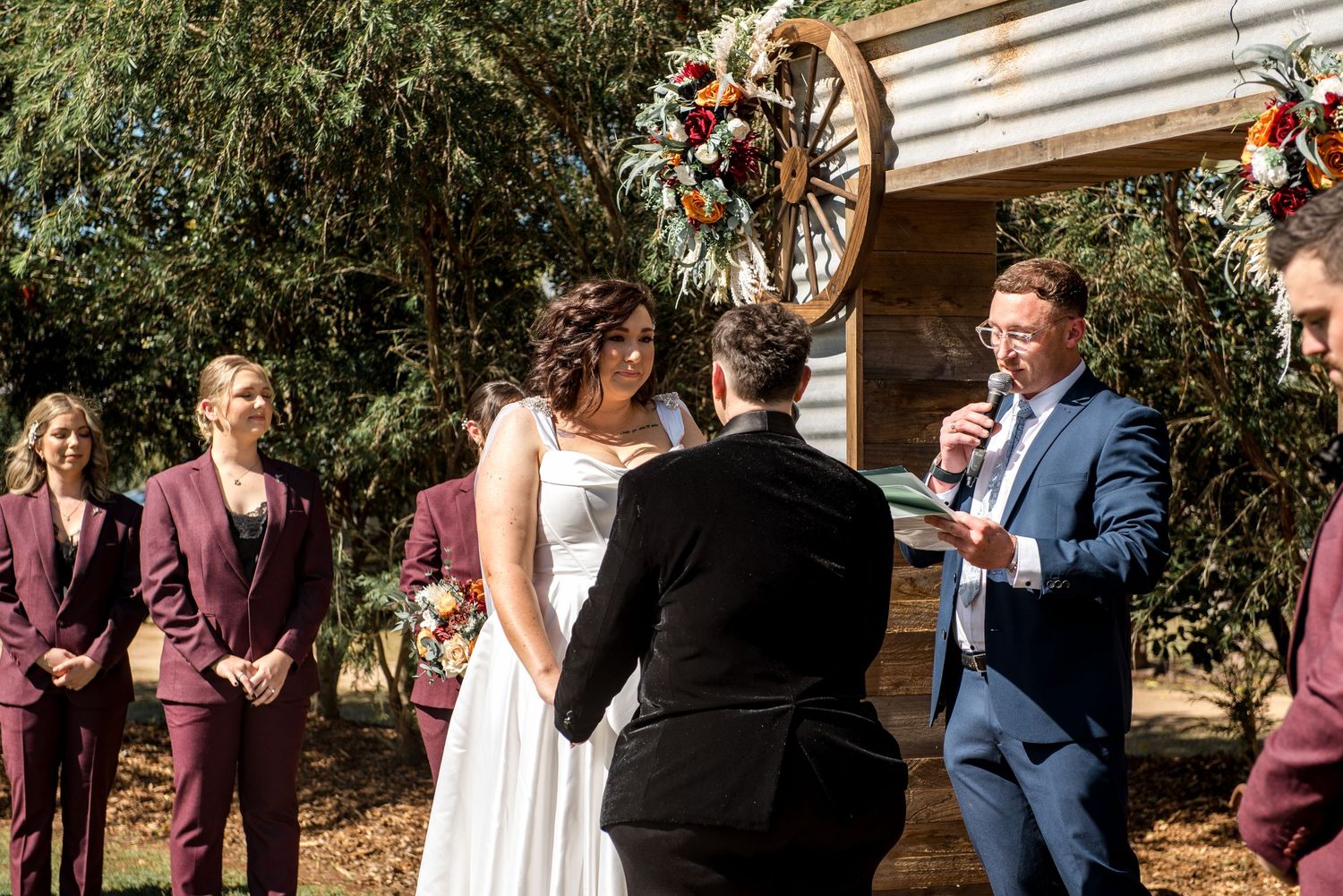 Smiling celebrant in blue suit reads vows as bride and groom hold hands at rustic outdoor wedding altar.