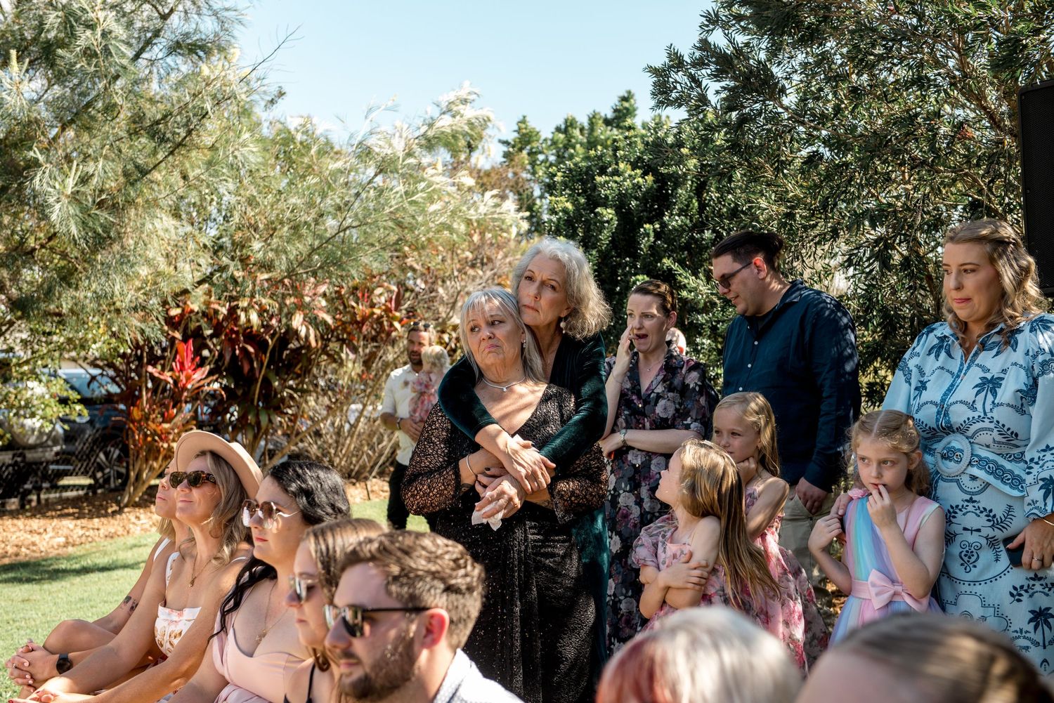 Wedding guests gathered outdoors in a garden, including elegantly dressed women and children enjoying the ceremony.