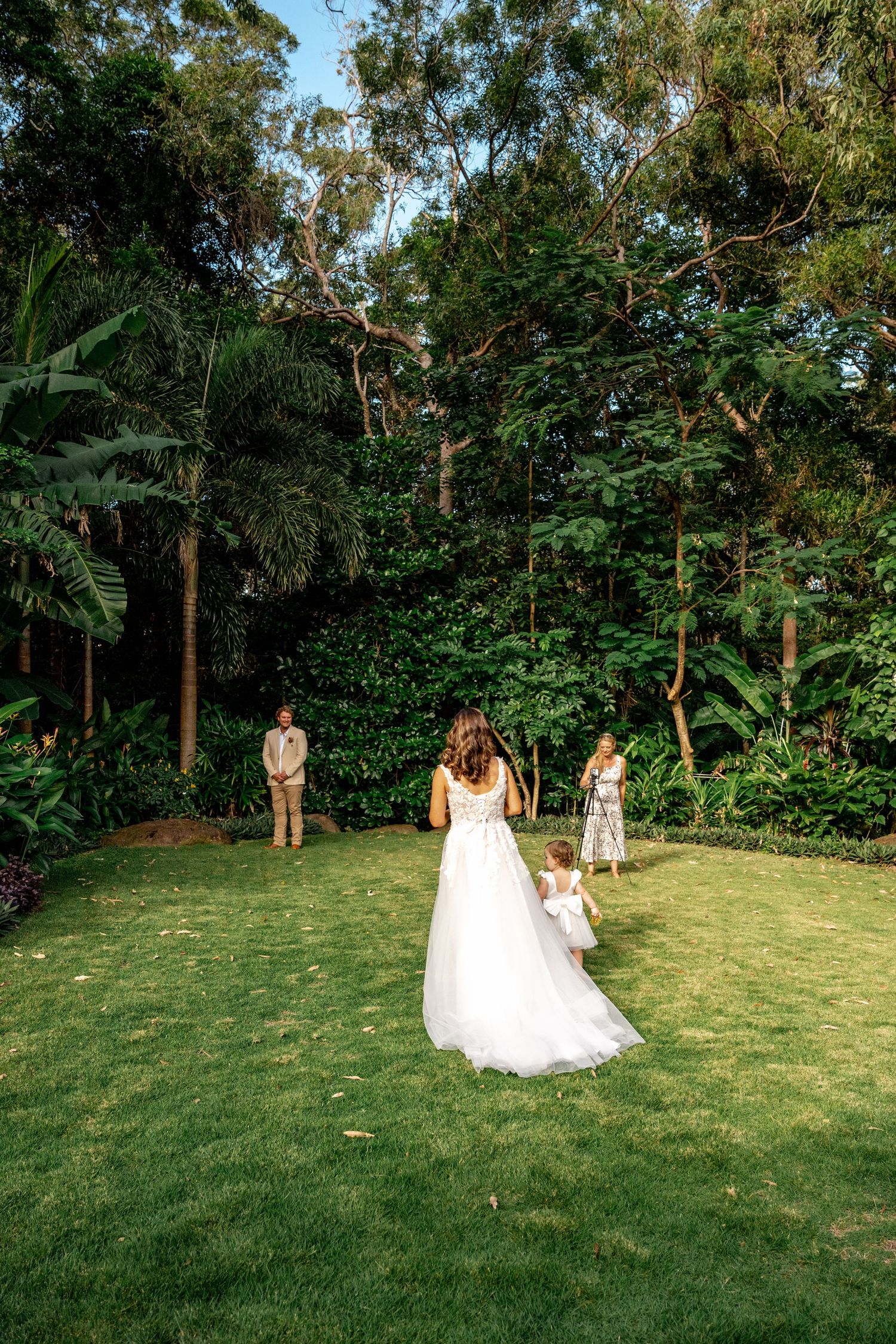 A bride in a white gown walks toward a groom with a child on a lush green garden lawn.