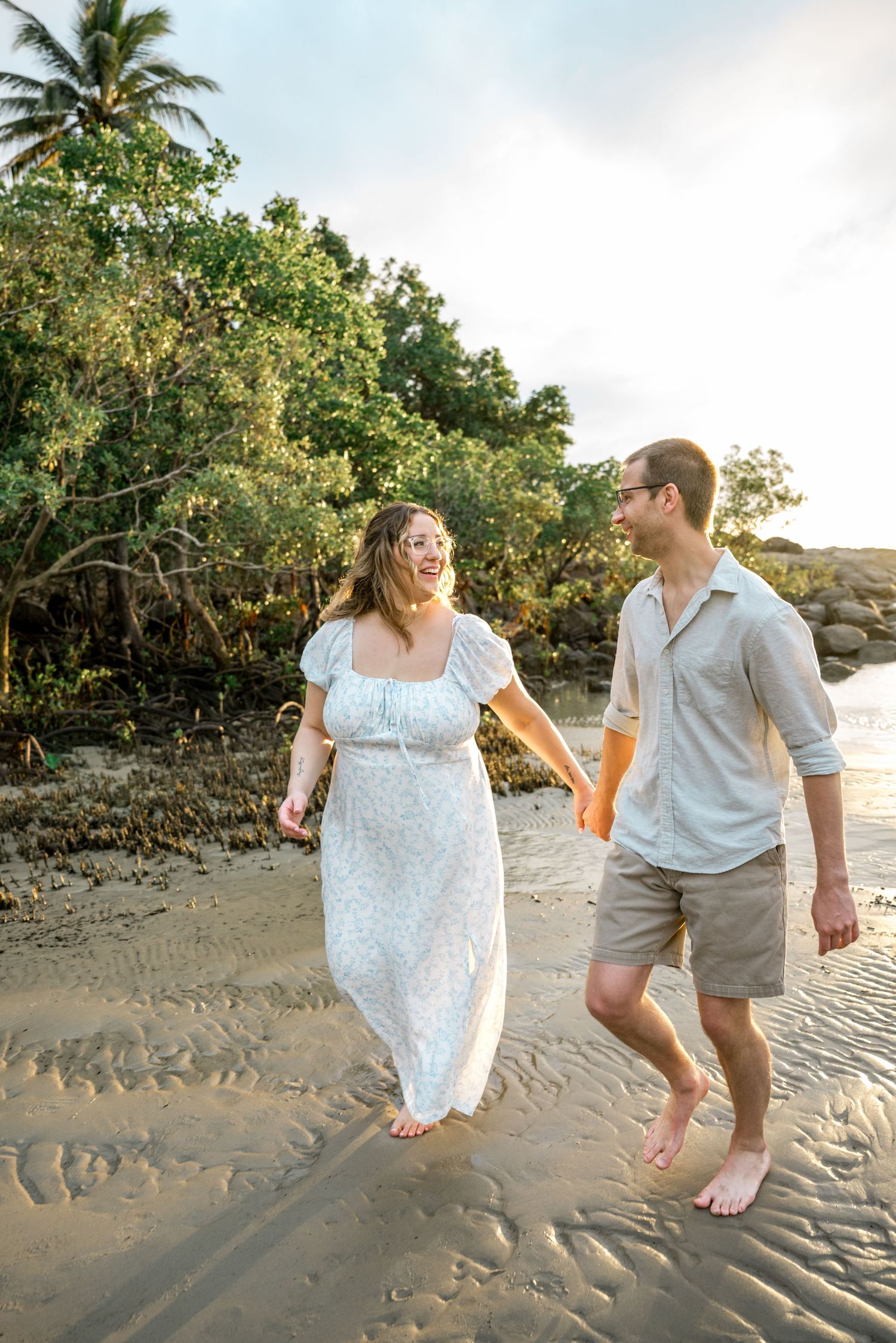 Laughing couple strolls barefoot on sandy tropical beach at sunset, woman in flowing white dress at Four Mile Beach, Port Douglas.