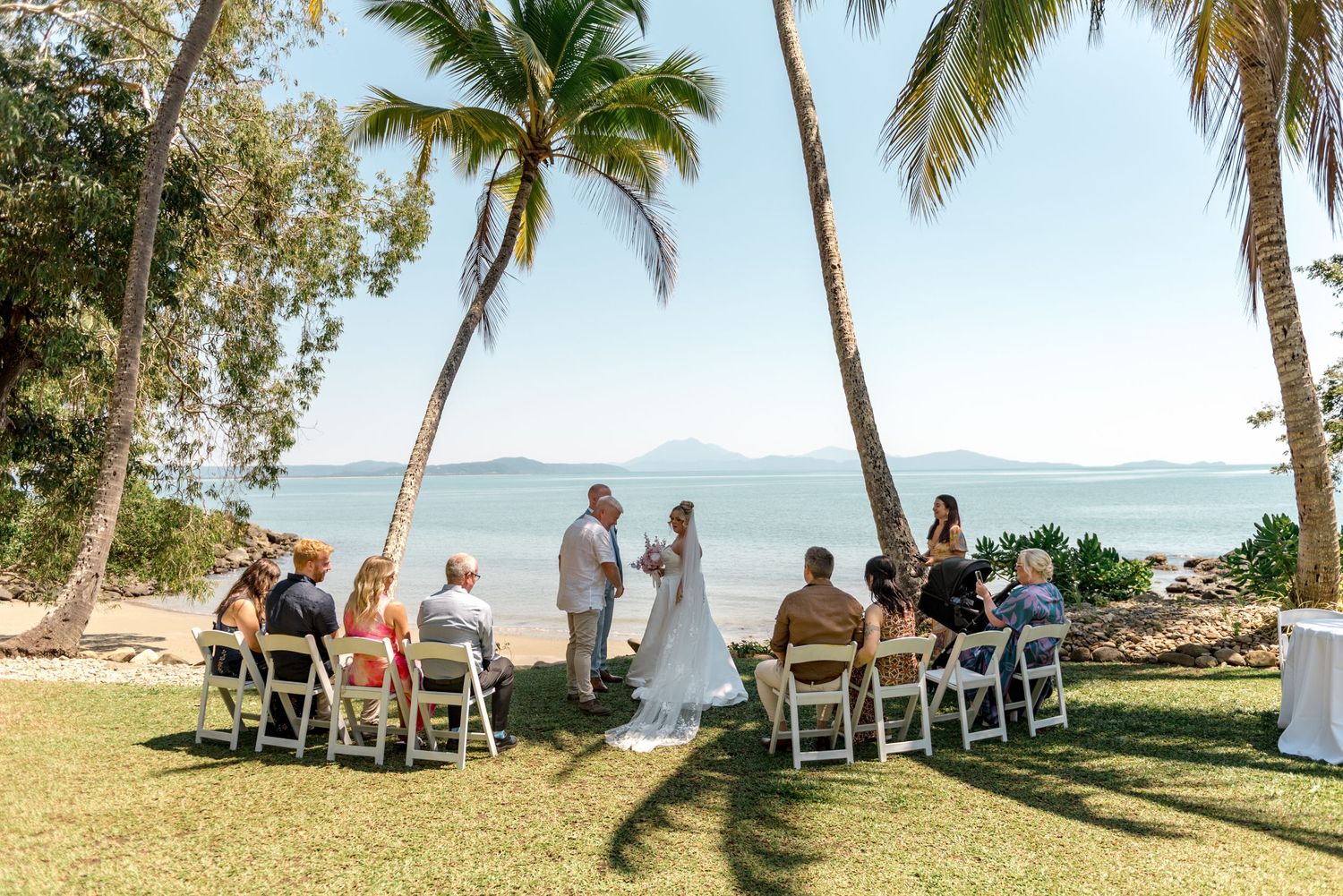 Small tropical beach wedding ceremony with guests seated on white chairs under tall palm trees.