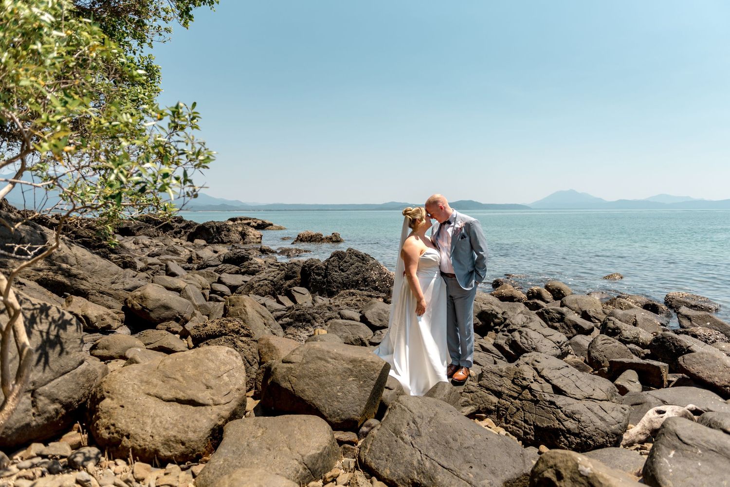 Wedding couple embraces on coastal rocks with stunning blue ocean and distant mountain views backdrop.