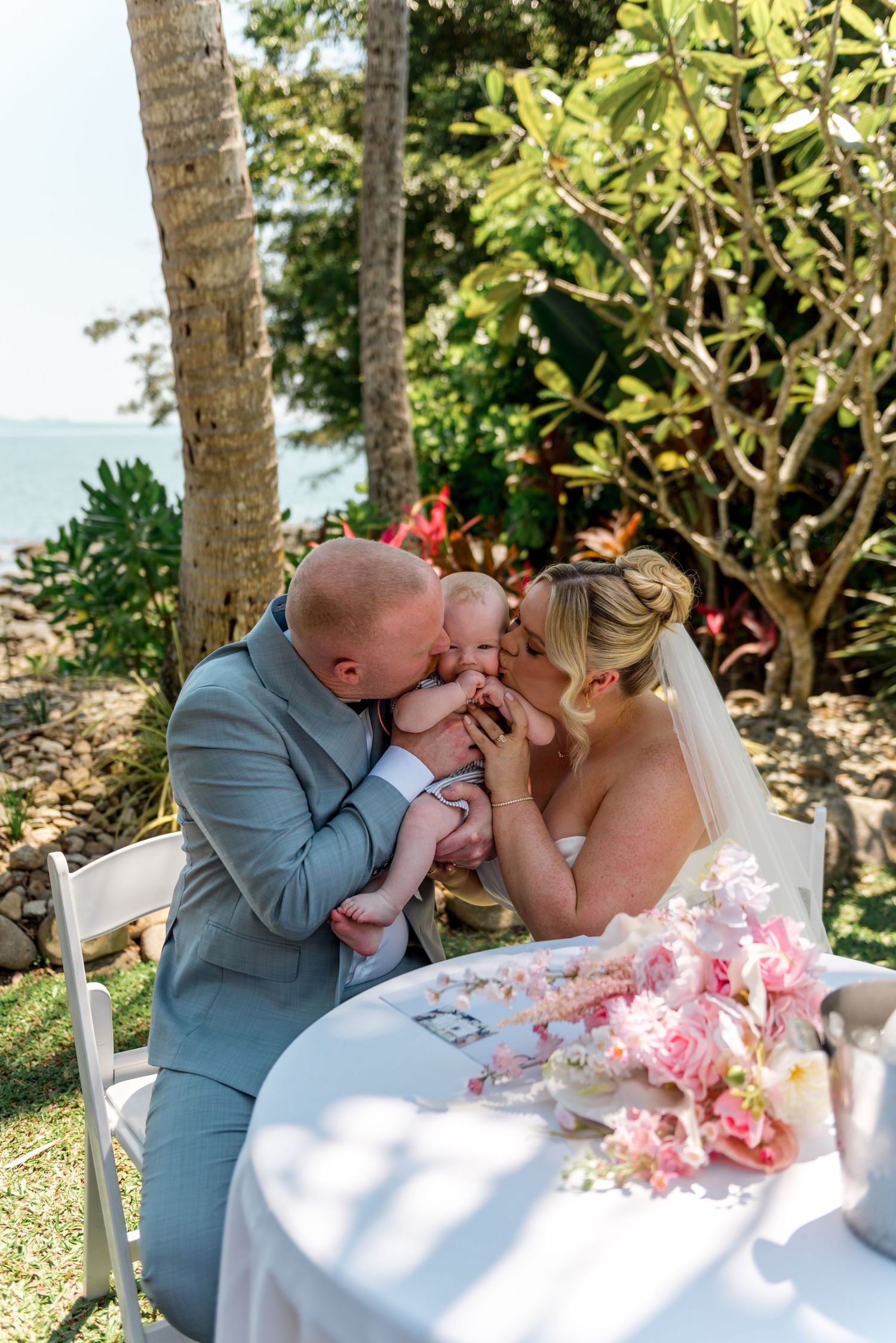 Bride and groom tenderly kissing their newborn baby at outdoor wedding table surrounded by tropical greenery.