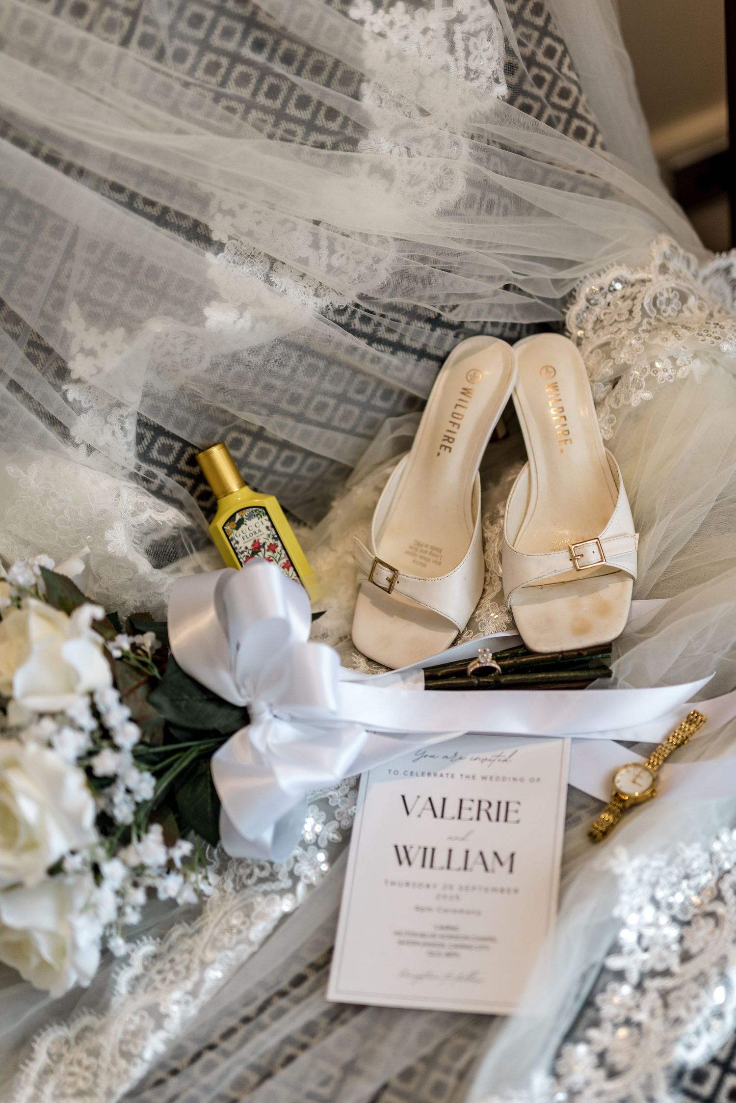 Wedding flat lay featuring white heels, invitation for Valerie and William, bouquet, perfume, and gold watch on lace veil.