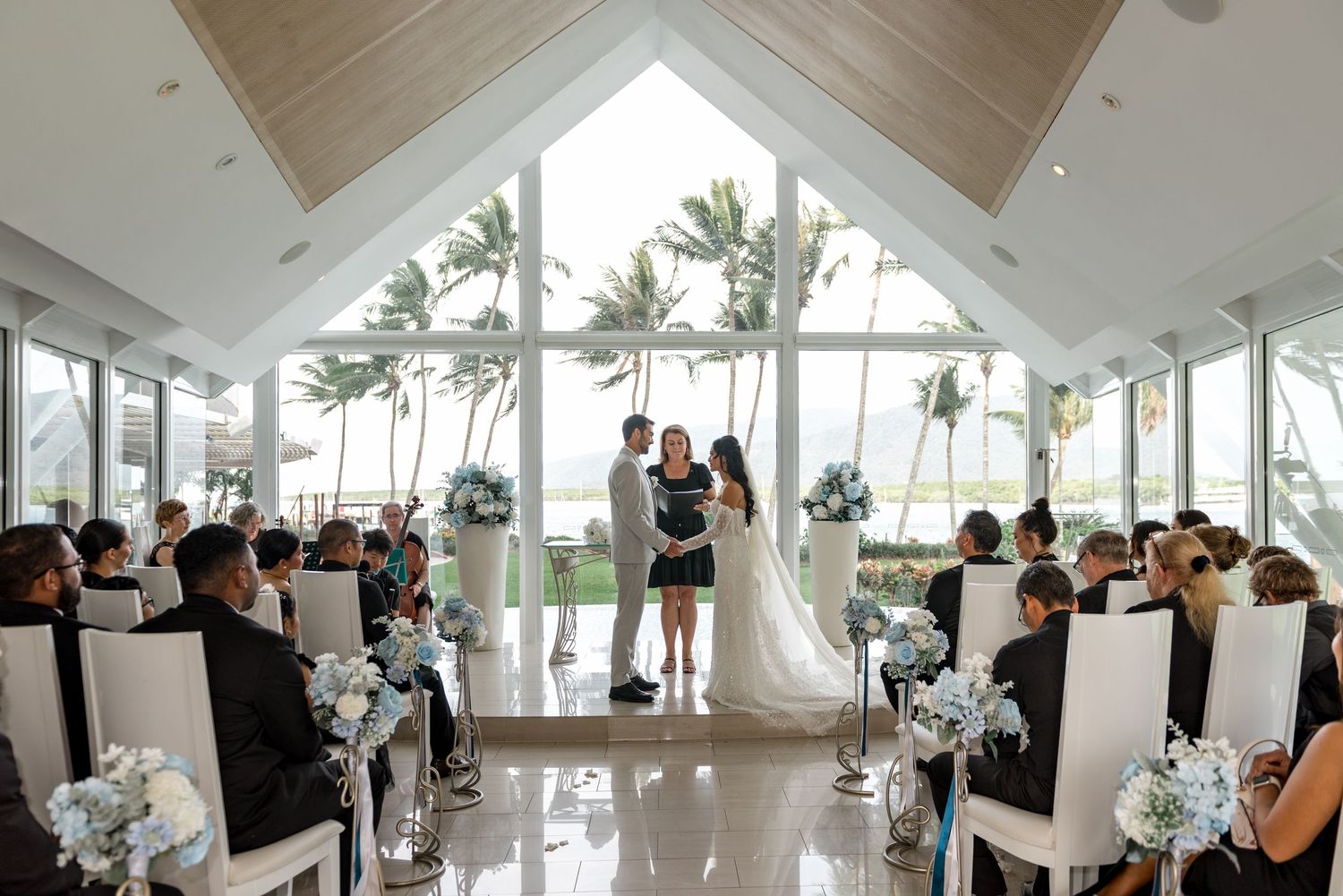 Wide angle view of wedding ceremony in bright glass chapel with palm tree backdrop.