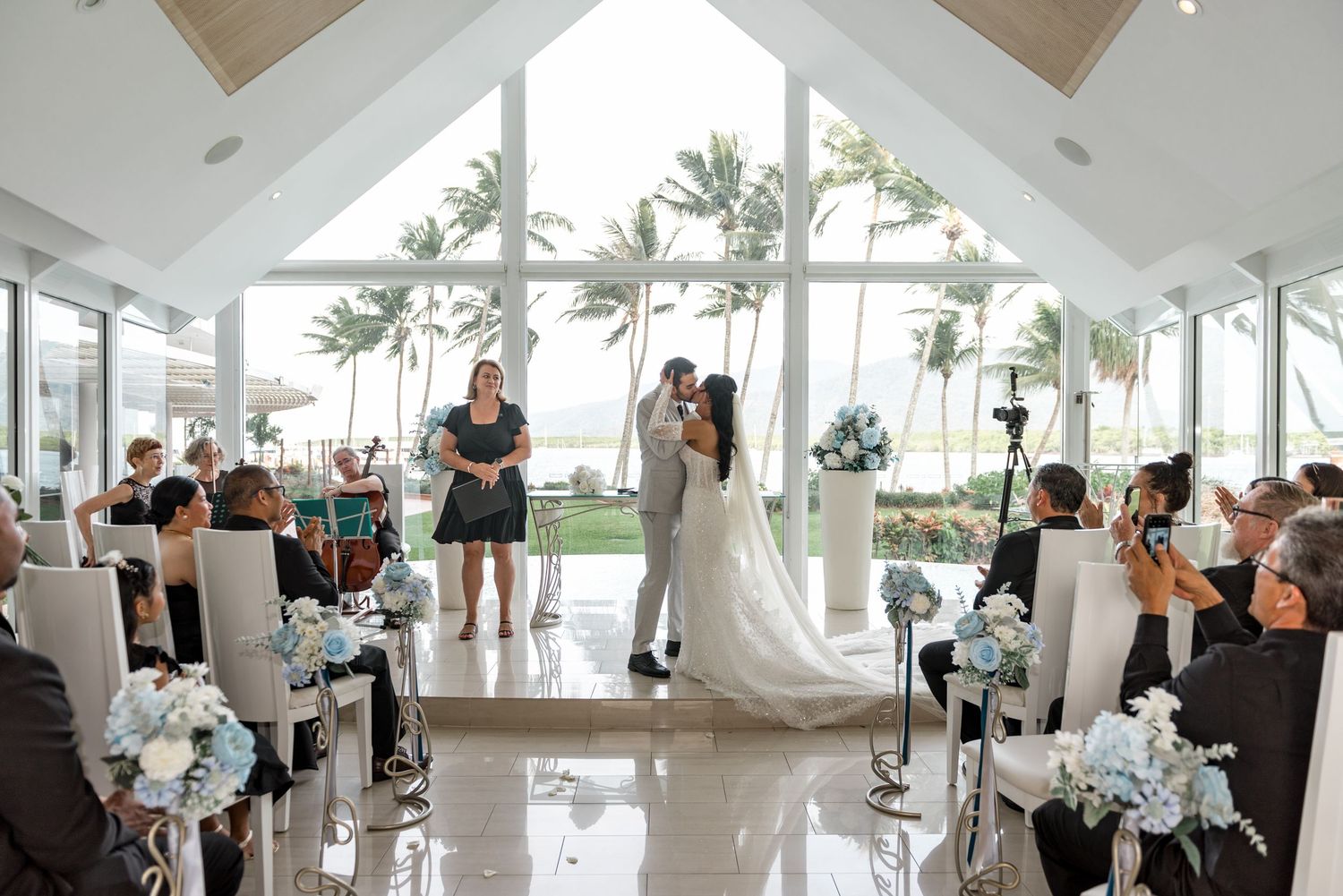 Elegant beach wedding ceremony in a modern chapel with floor-to-ceiling windows, palm trees, and blue floral arrangements.