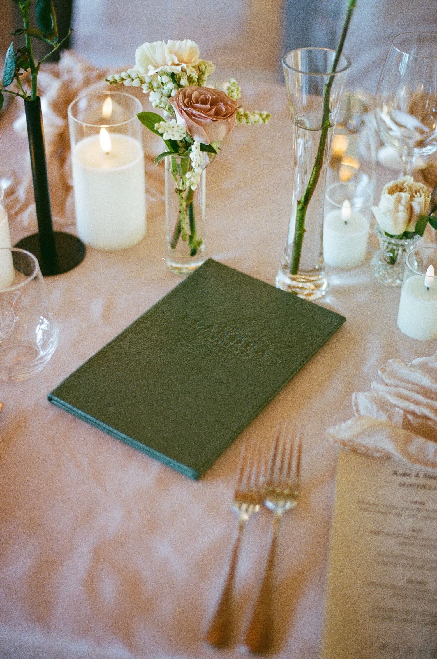 Wedding table setting with green menu book, pink roses, white candles, and delicate floral bud vases.