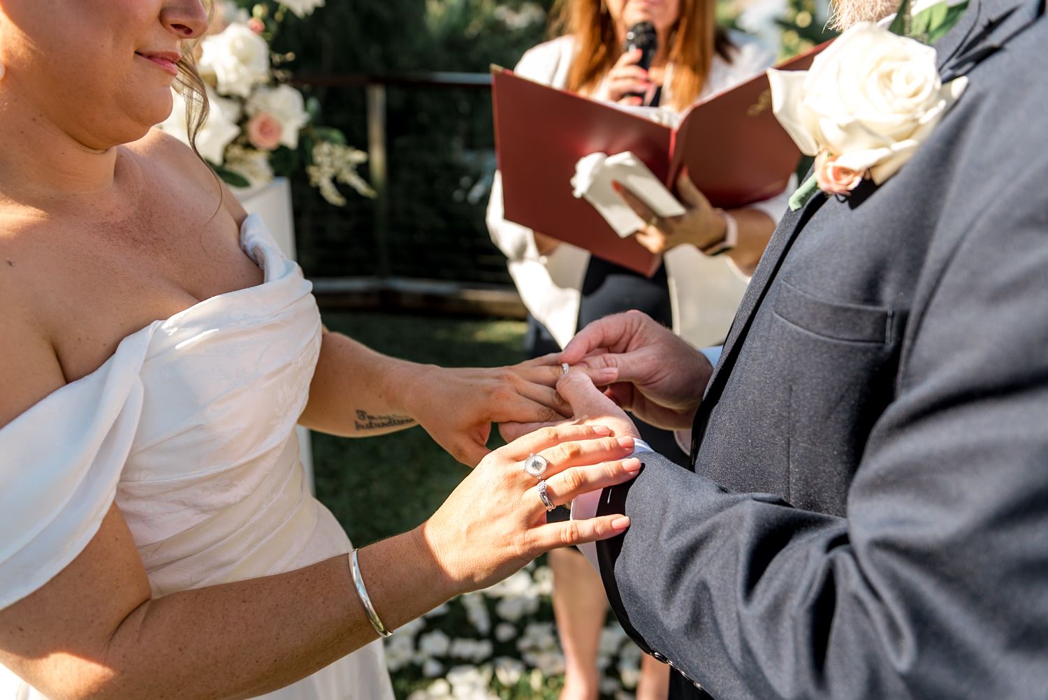 Groom places wedding ring on brides finger during outdoor ceremony as officiant reads from red book.