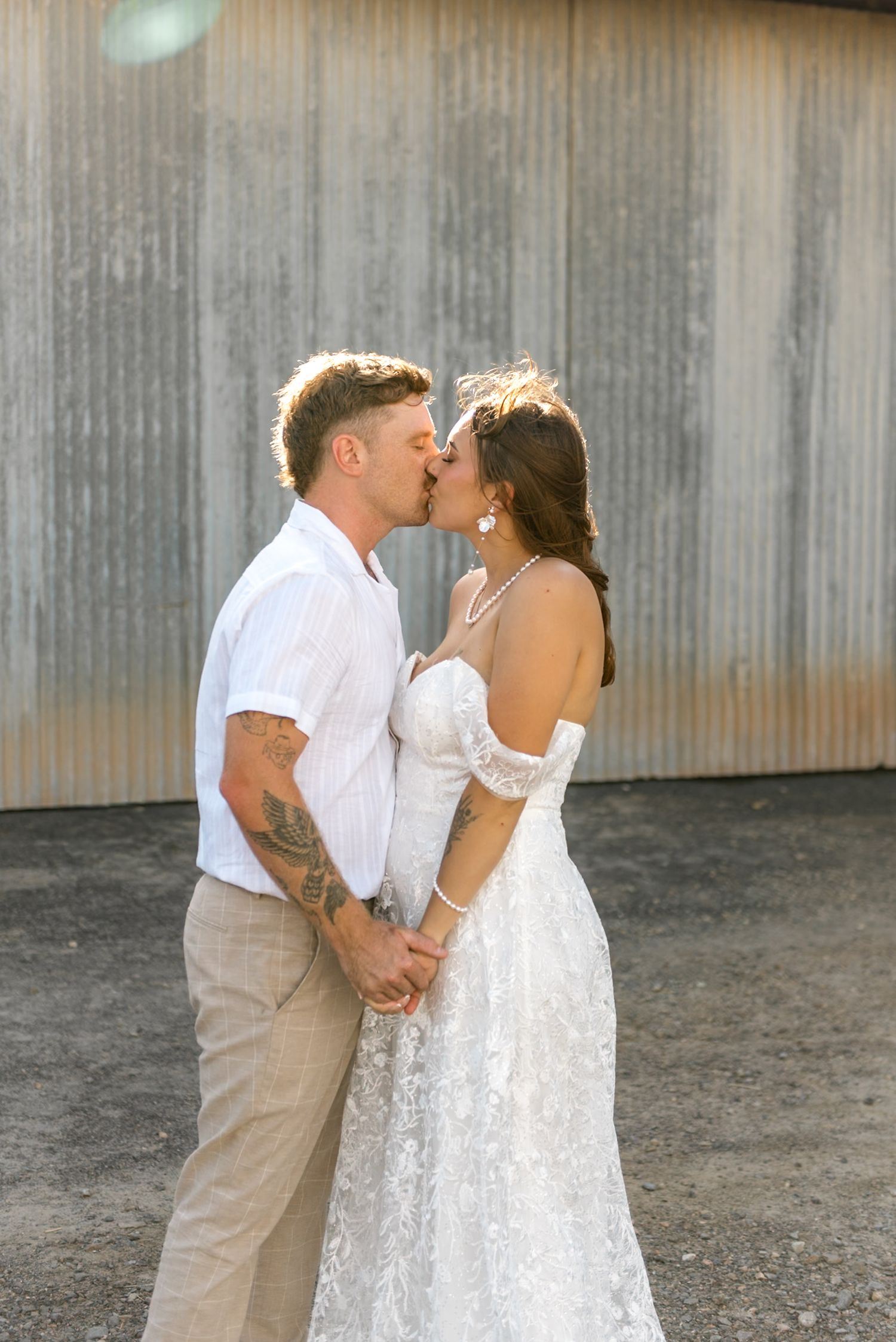 Bride and groom share a romantic kiss while holding hands in golden sunlight before a corrugated metal wall.