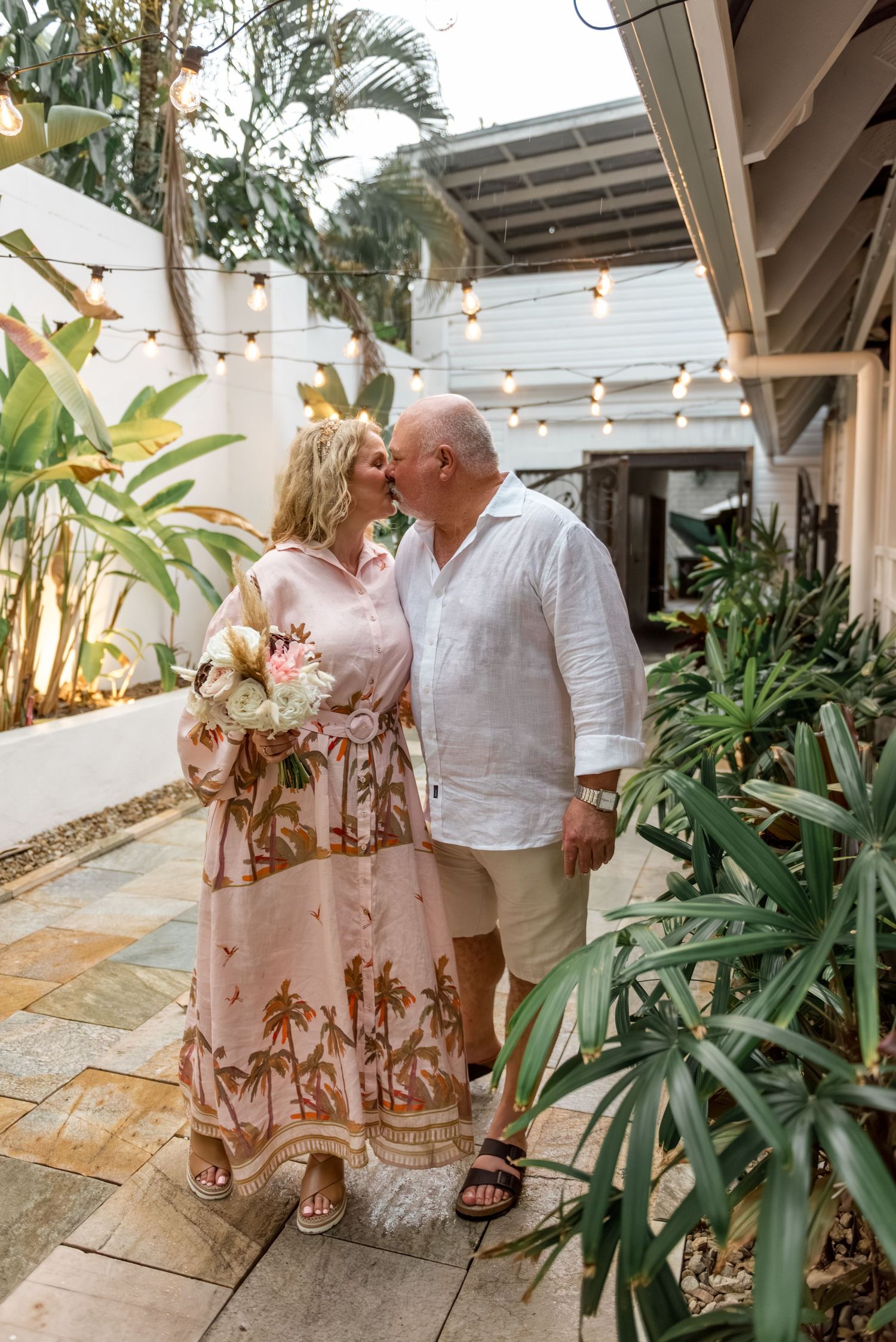 Couple sharing a romantic kiss in a tropical garden courtyard with string lights.